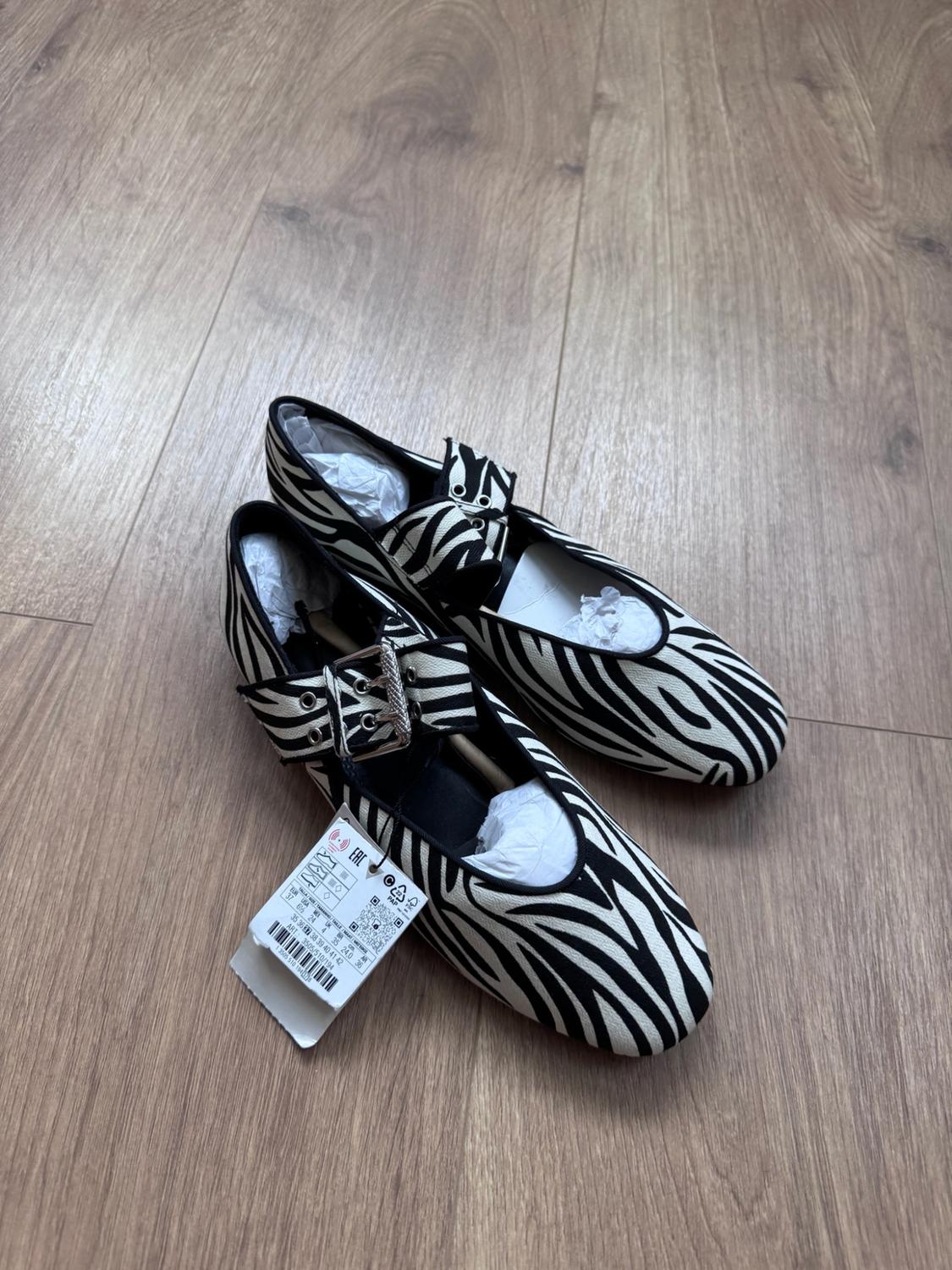 Flat shoes with a black-and-white zebra print, strap and silver buckle. They have a label, on a wooden floor.