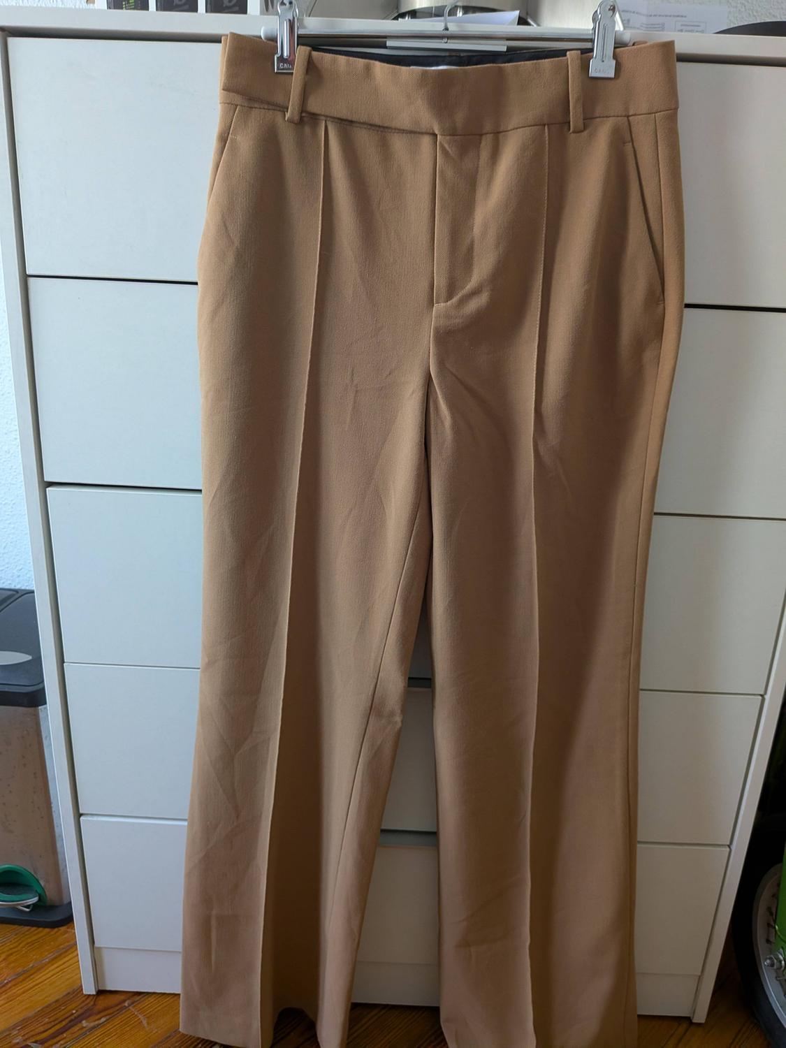 Camel-coloured dress trousers hanging on a hanger in front of a white chest of drawers on a wooden floor. Relaxed atmosphere.