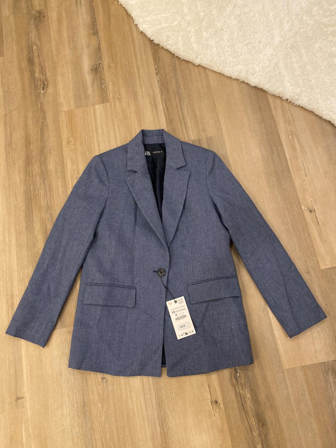 One-button blue jacket on light wood flooring, with visible hang tag. Part of a white rug in the upper corner.