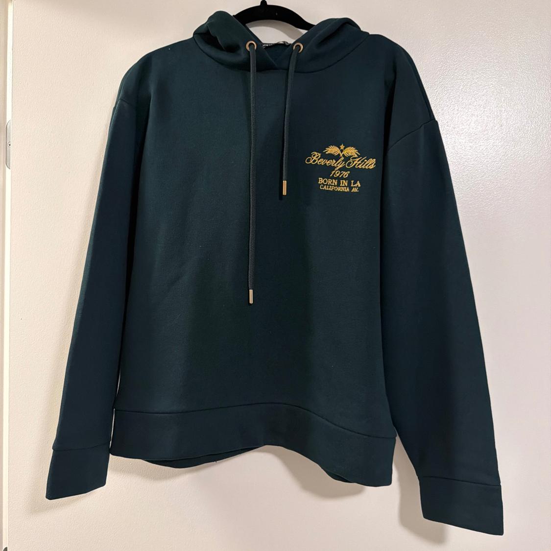 Dark green hooded sweatshirt hanging against a white background. Embroidered text: Beverly Hills 1976 BORN IN LA CALIFORNIA AV.