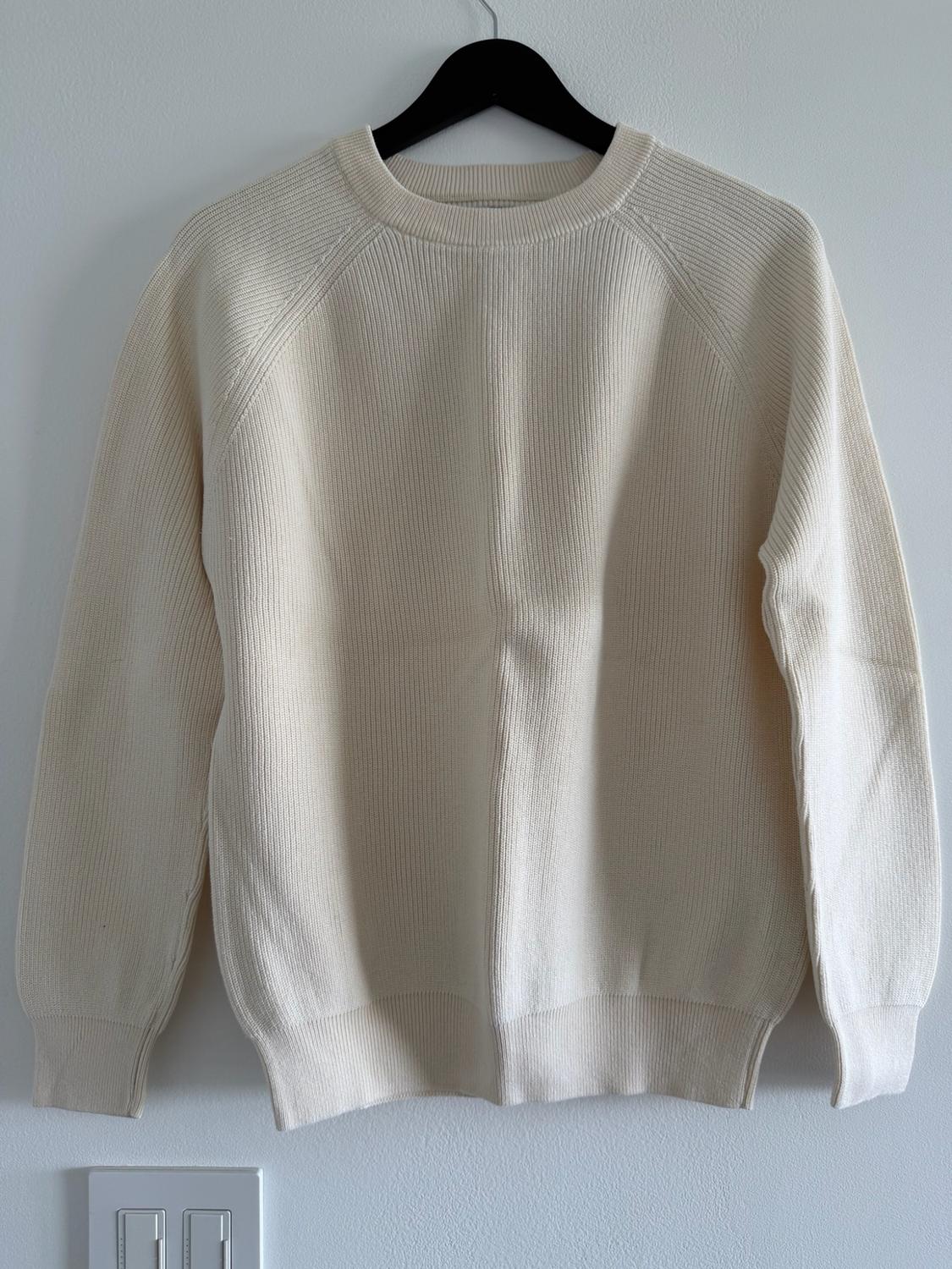 Cream-colored ribbed long-sleeve knit sweater hanging on a black hanger against a white wall.
