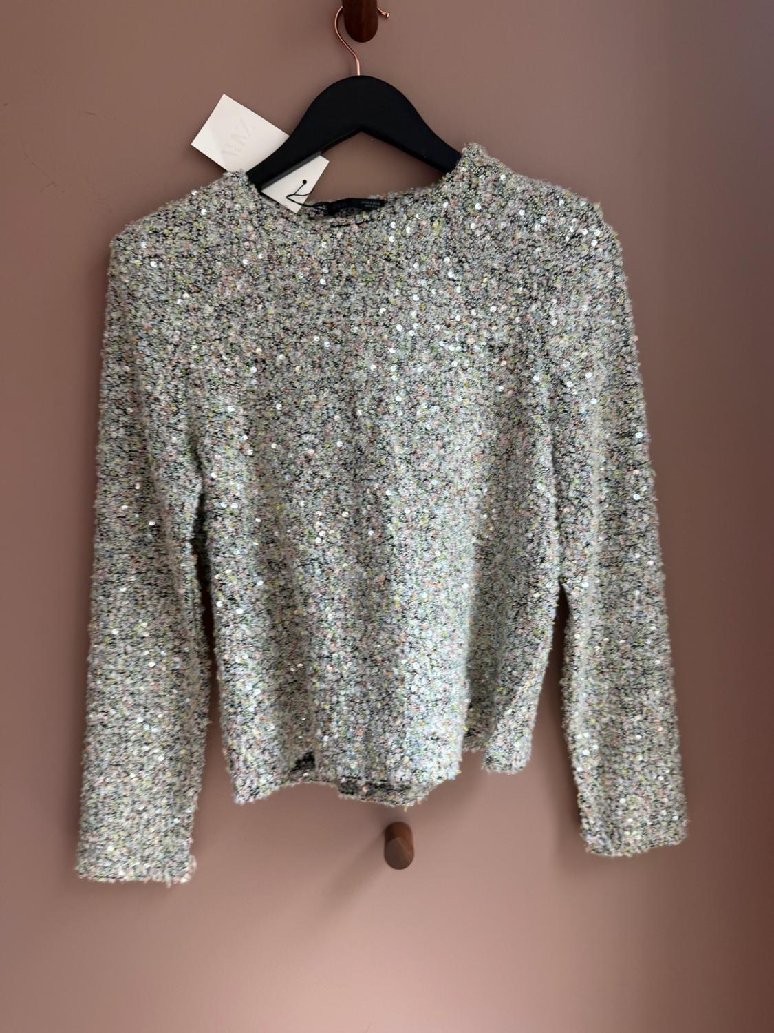 Light-coloured long-sleeved jumper with texture and sequins, hanging on a black hanger against a pink wall.