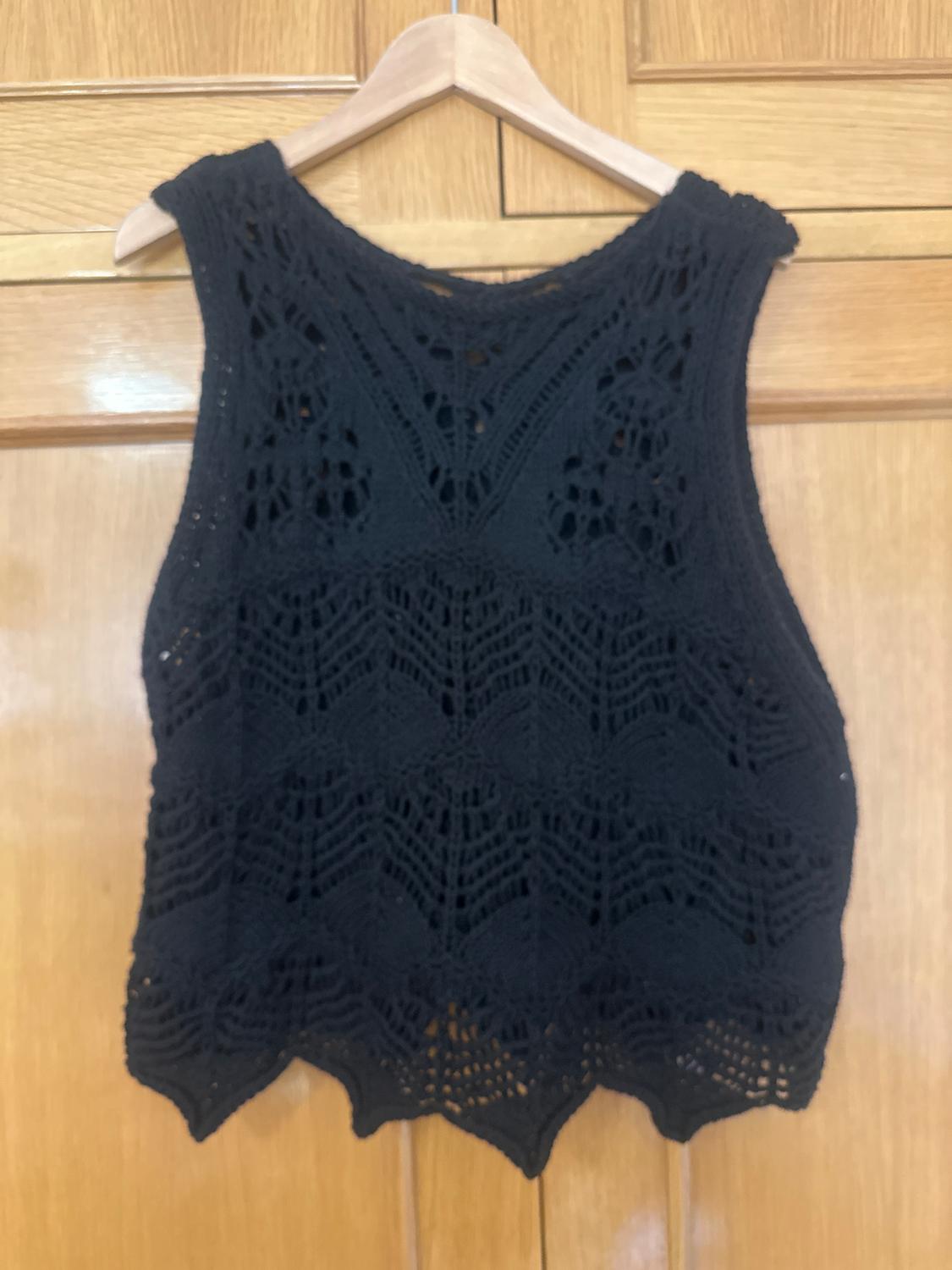 Black crochet waistcoat hanging on a wooden hanger over a light wooden door. Design with geometric patterns.