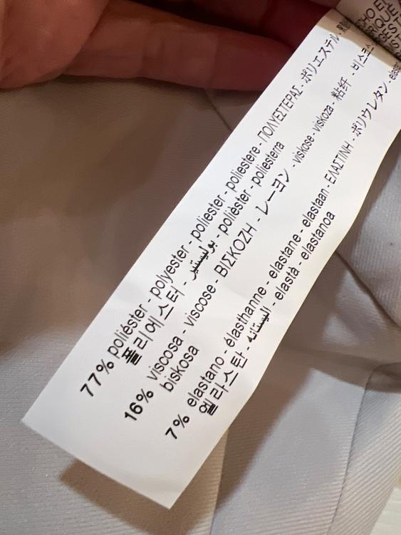 White fabric label with composition: 77% polyester, 16% viscose, 7% elastane, in various languages. Beige fabric background.