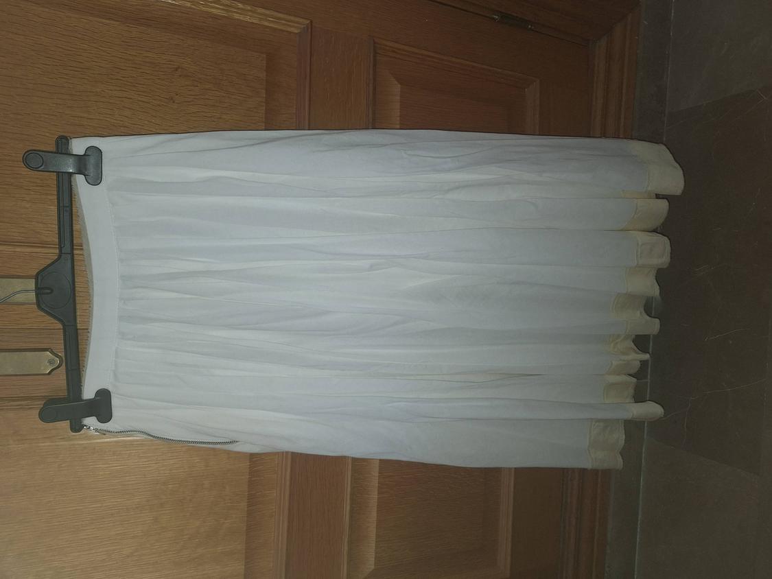 White pleated skirt on a black hanger, hanging in front of a wooden door. Features a cream-toned hem and side zip.