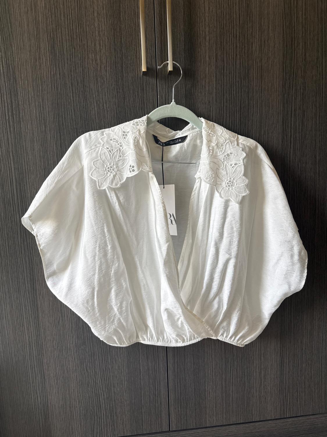 White wrap blouse with an elasticated hem and embroidered floral collar, hanging in a dark wood wardrobe. ZARA label.