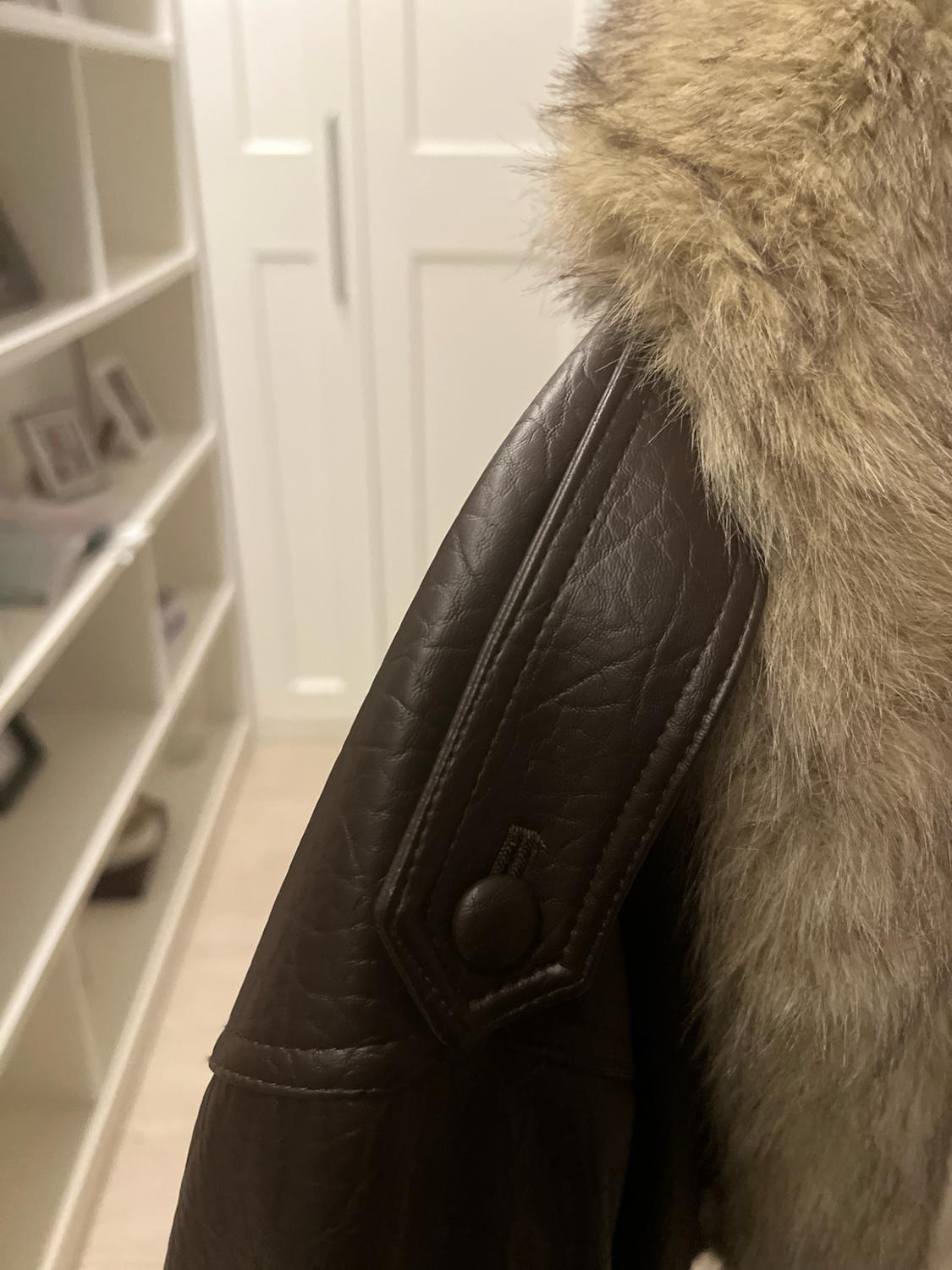 Close-up of a textured brown jacket with a fur collar. Shoulder detail with a strap and button. Blurred white wardrobe background.