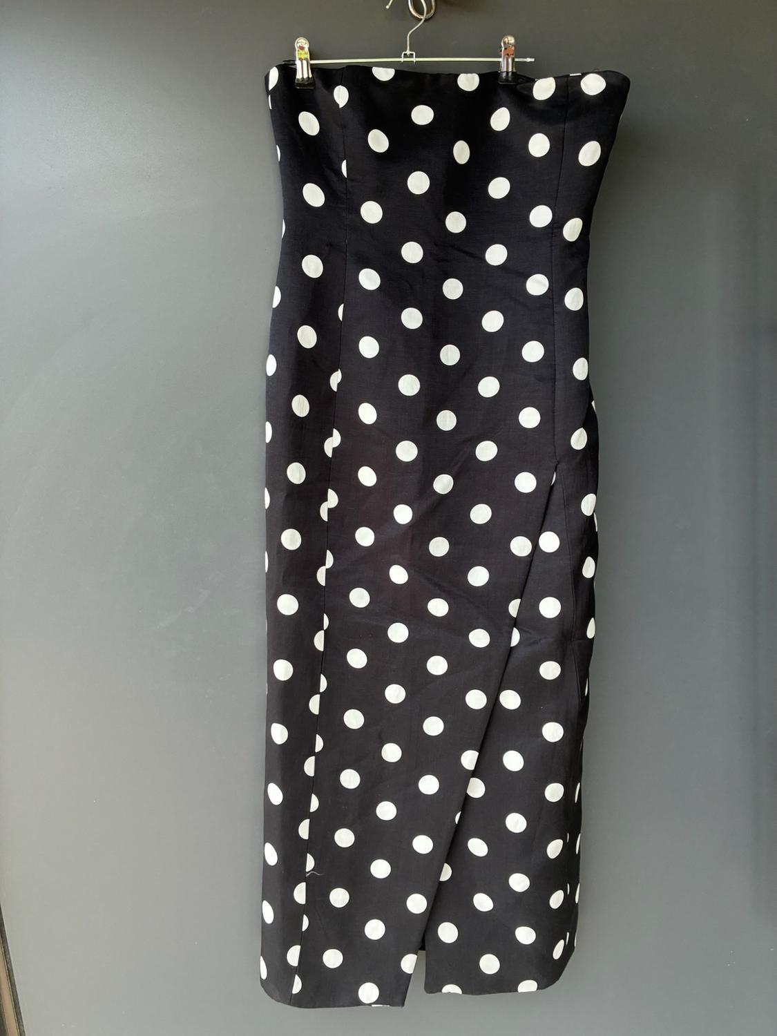 Black midi dress with white polka dots and a bandeau neckline, hanging from a metal hanger against a grey wall.