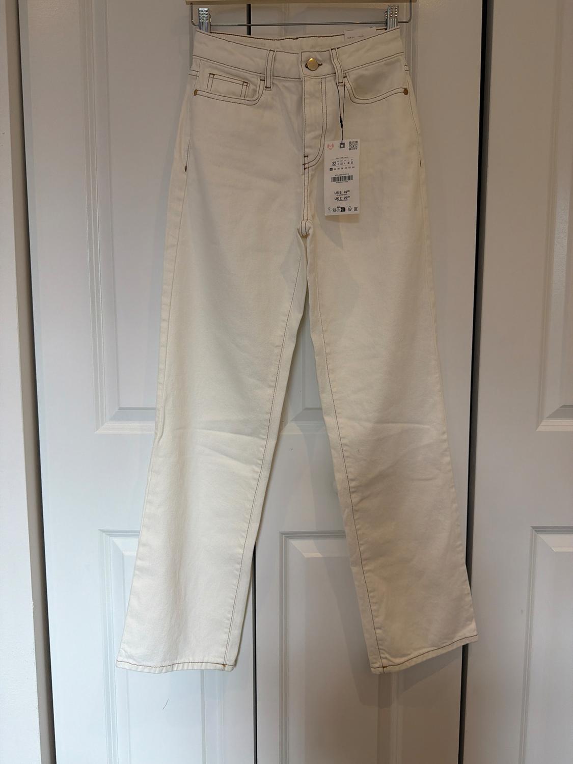 Ecru straight-leg jeans with contrast stitching and a tag, hanging on a clear hanger in front of a white door.