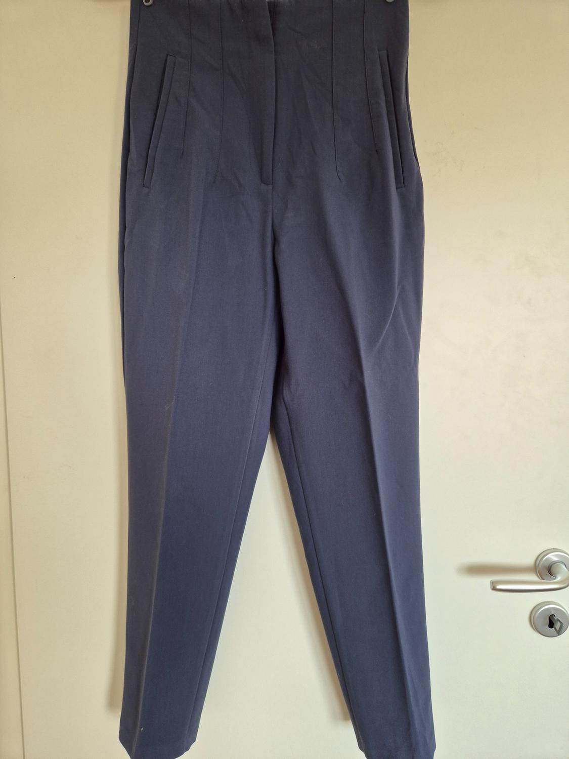Dark blue trousers hanging on a white door, with pleats and side pockets. Neutral and minimalist background.