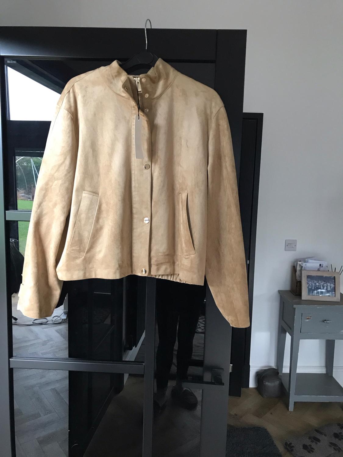 Beige jacket with a tag hanging on a black door. A person is reflected in the glass. On the right, a grey table in an interior.