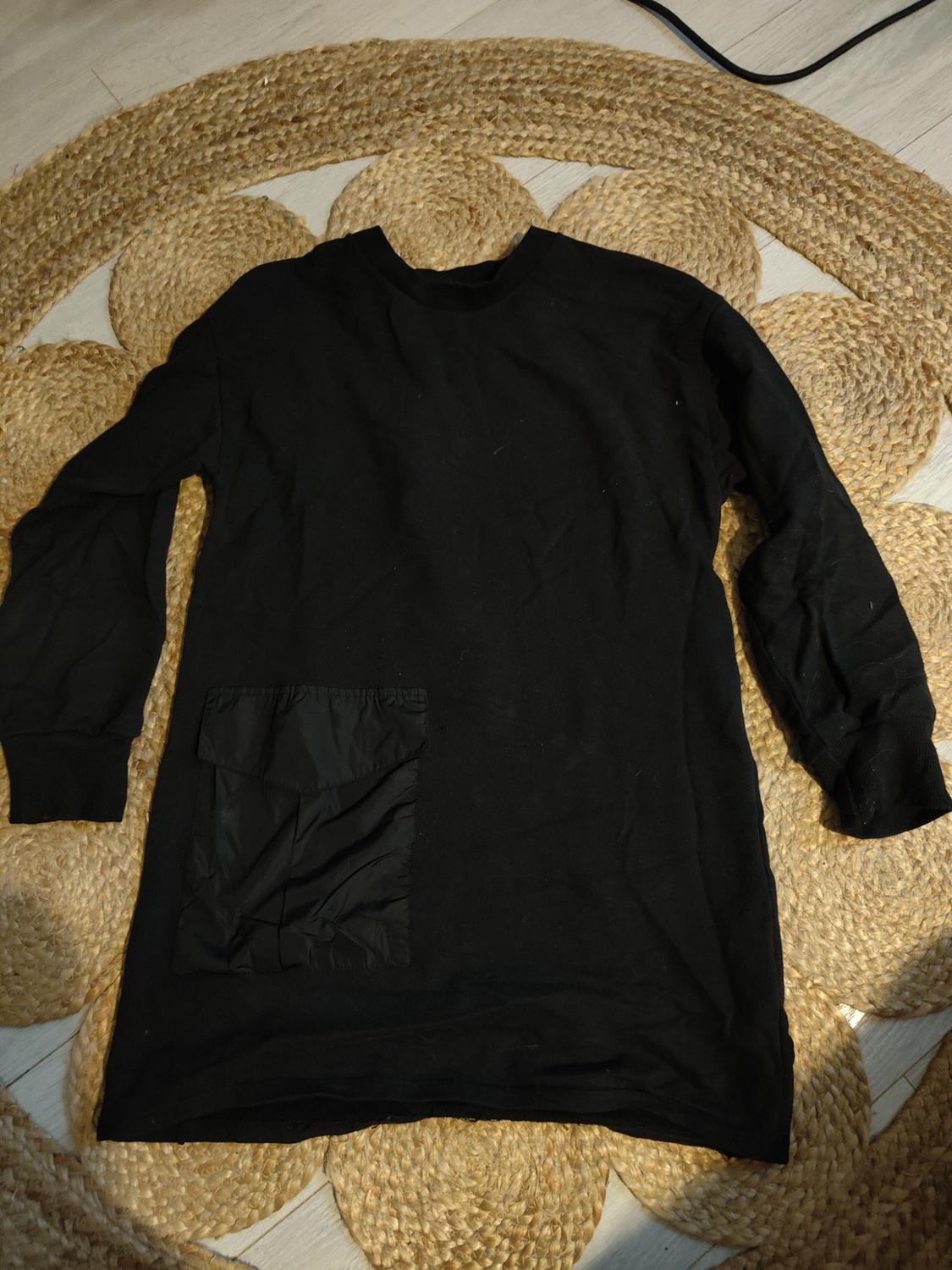 Black long-sleeved sweatshirt with a different fabric pocket on the bottom left, on a braided jute rug.