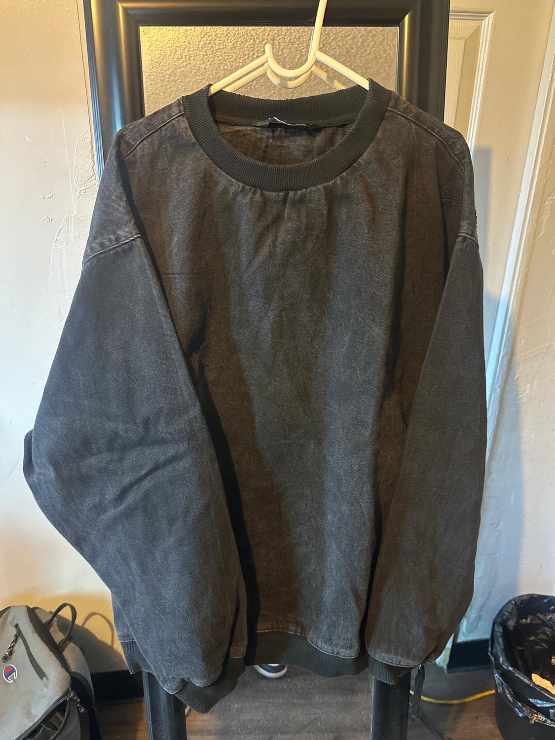 Dark grey sweatshirt hanging on a white hanger against a light background. Bottom of a backpack visible in the corner.