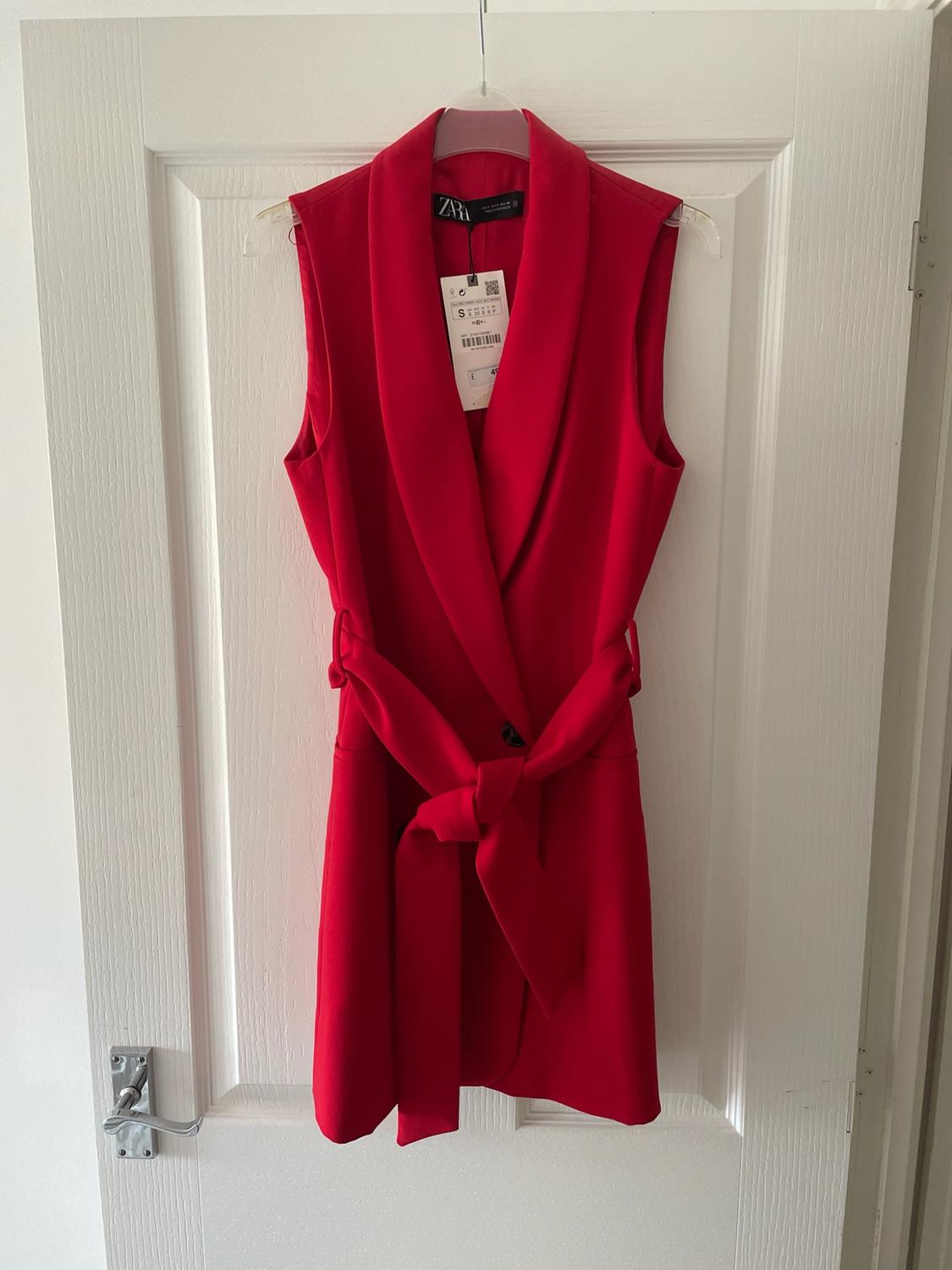 Long red waistcoat with lapels and a tied belt, hanging on a white door. Text: ZARA.