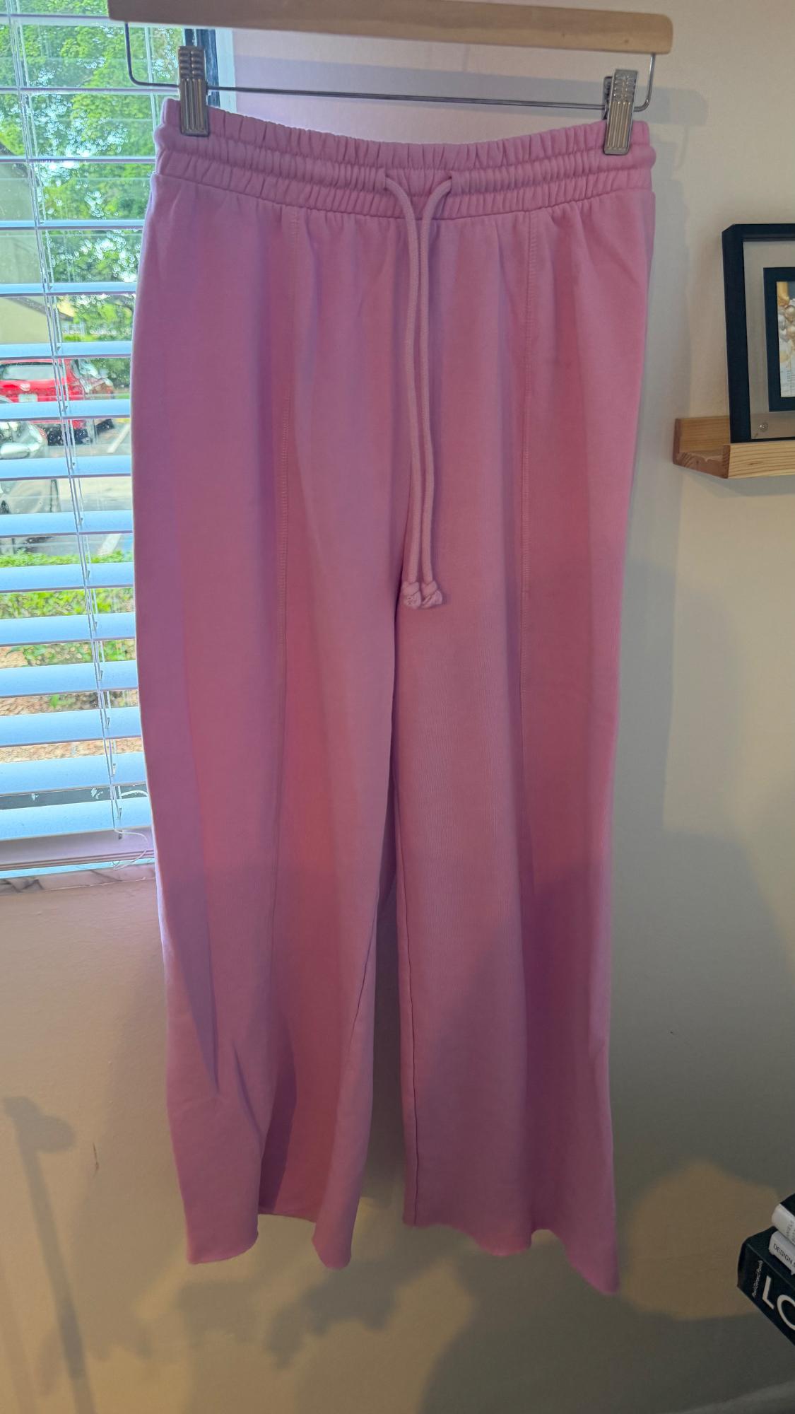 Wide-leg pink trousers with an elasticated drawstring waist, hanging on a hanger next to a window with white blinds.