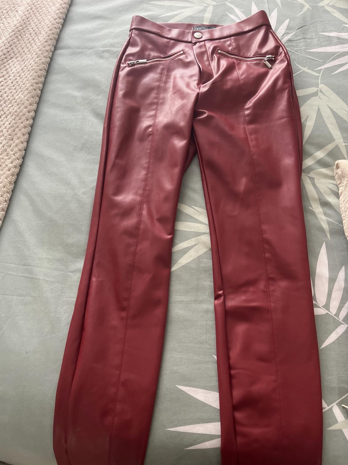 Red synthetic trousers with silver zips on the pockets, on a bed with a green and beige quilt.