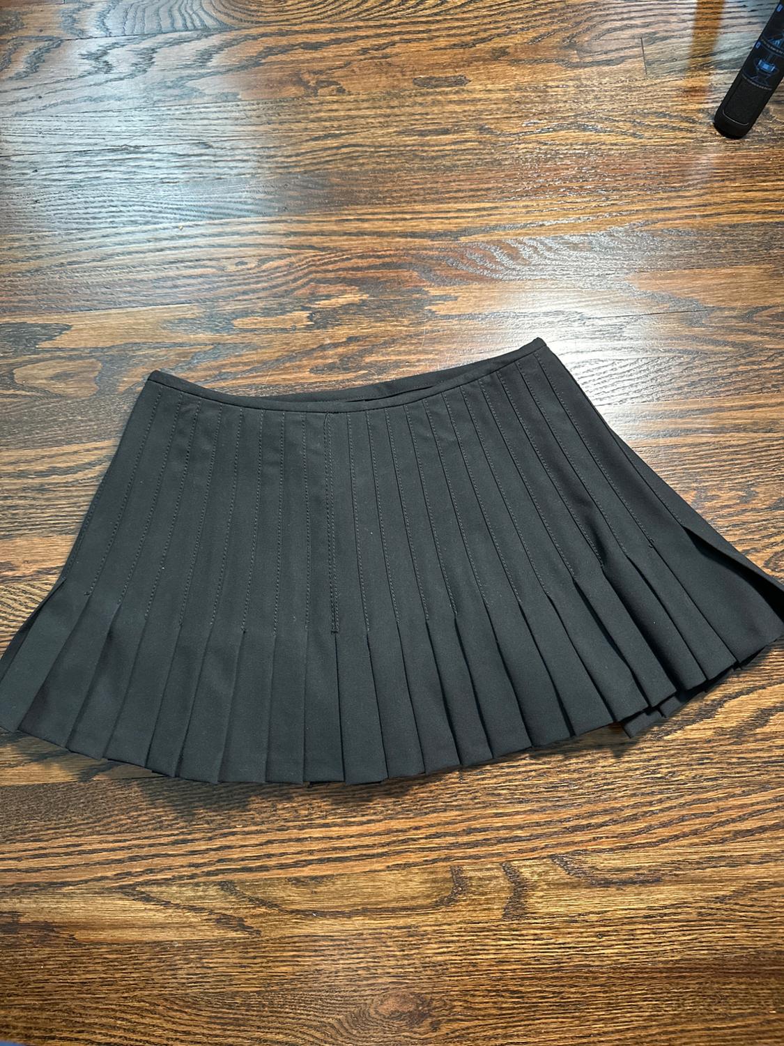 Black pleated skirt on a wooden floor with visible grain. The skirt has a classic and elegant design.