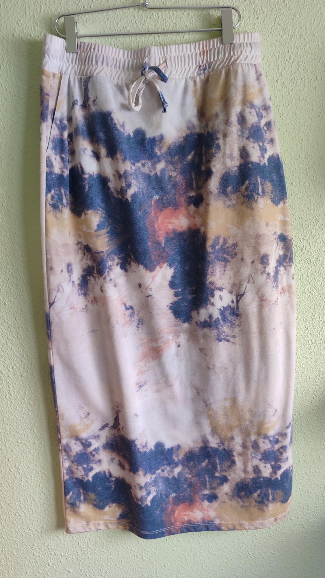 Skirt with tie-dye print in shades of blue, white and pink. Elasticated waist with drawstring, hanging on a hanger against a light green wall.