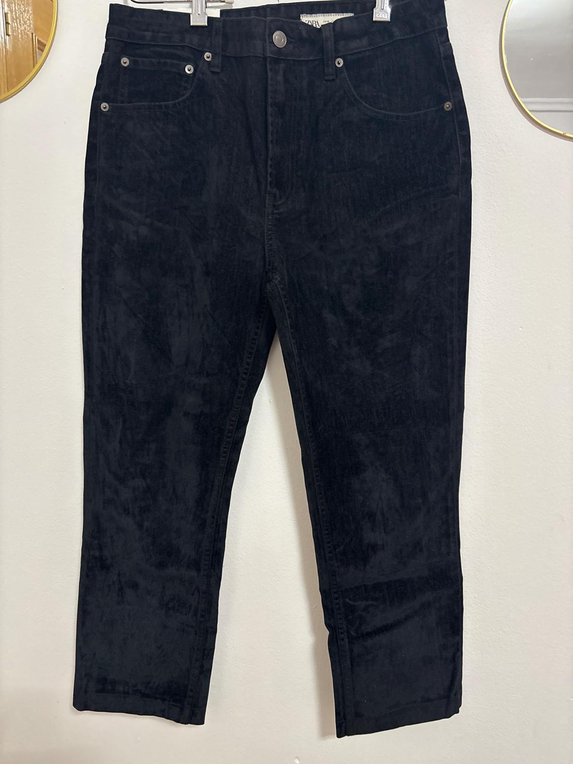 Black corduroy trousers hanging on a clothes hanger, white background with details of circular gold mirrors visible on the edges.