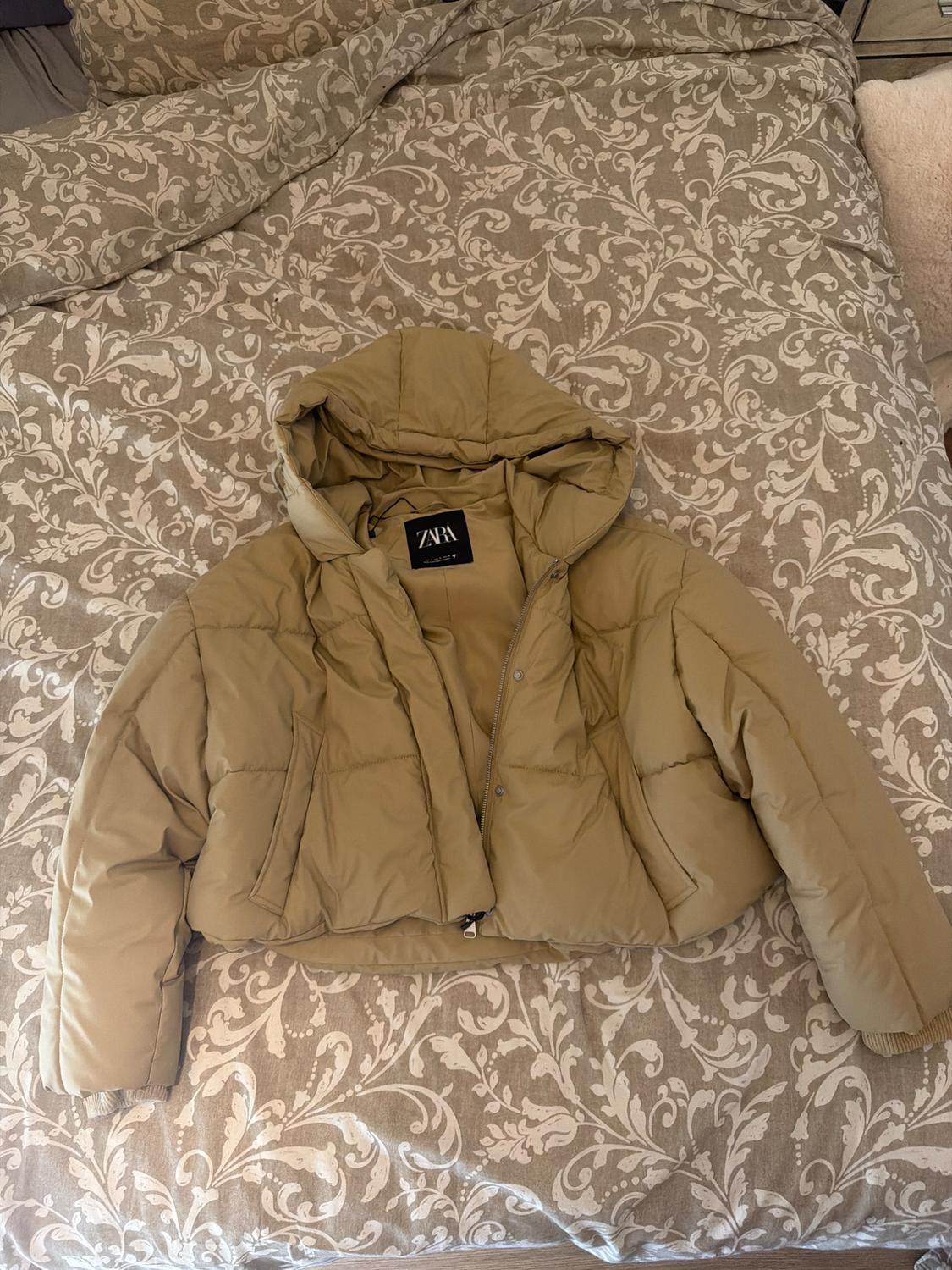 Beige padded jacket with a hood on a bed with a floral-patterned bedspread. Label bearing the text ZARA.