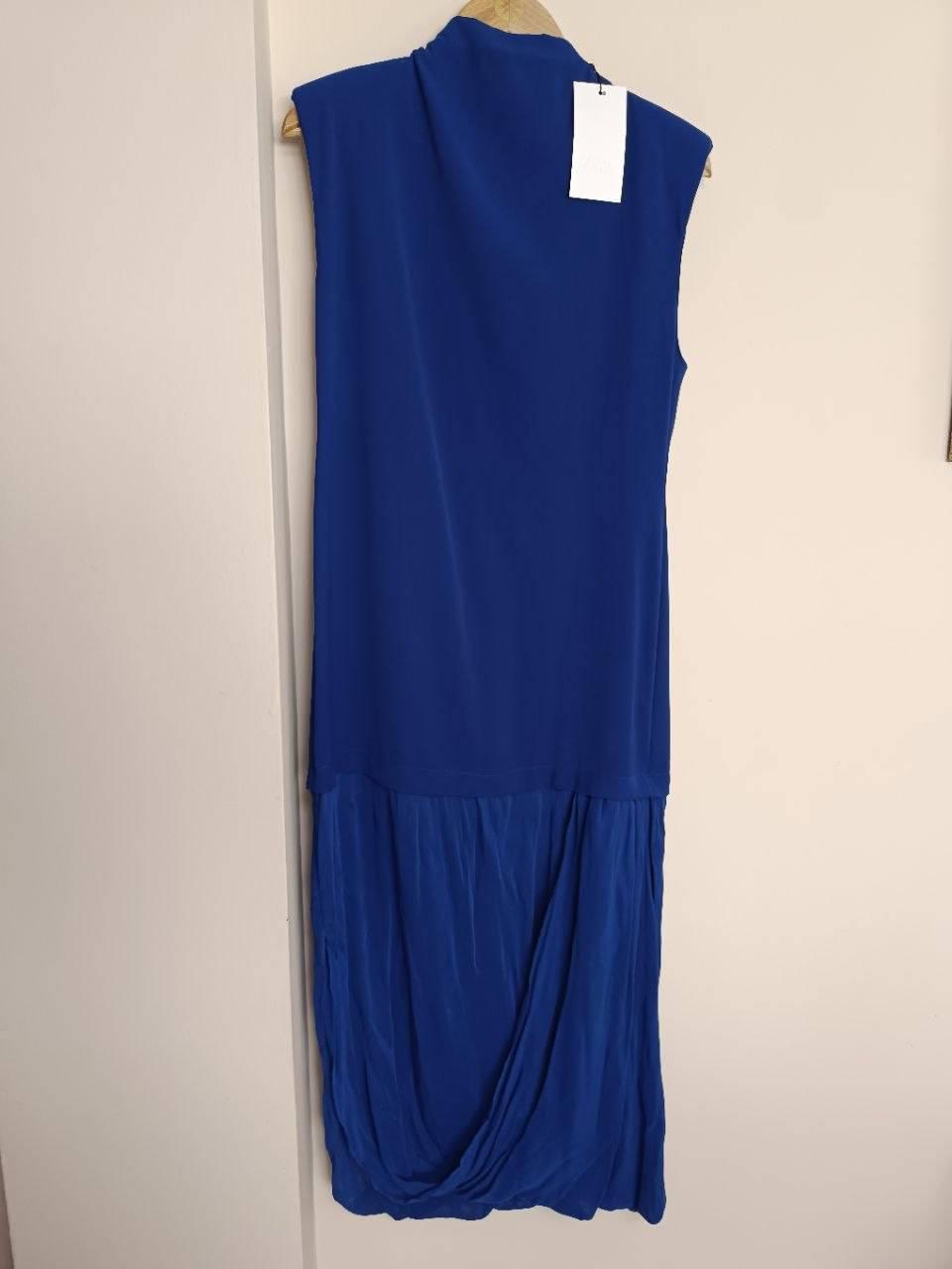 Sleeveless long blue dress with a draped skirt, hanging on a wooden hanger in front of a white wall.