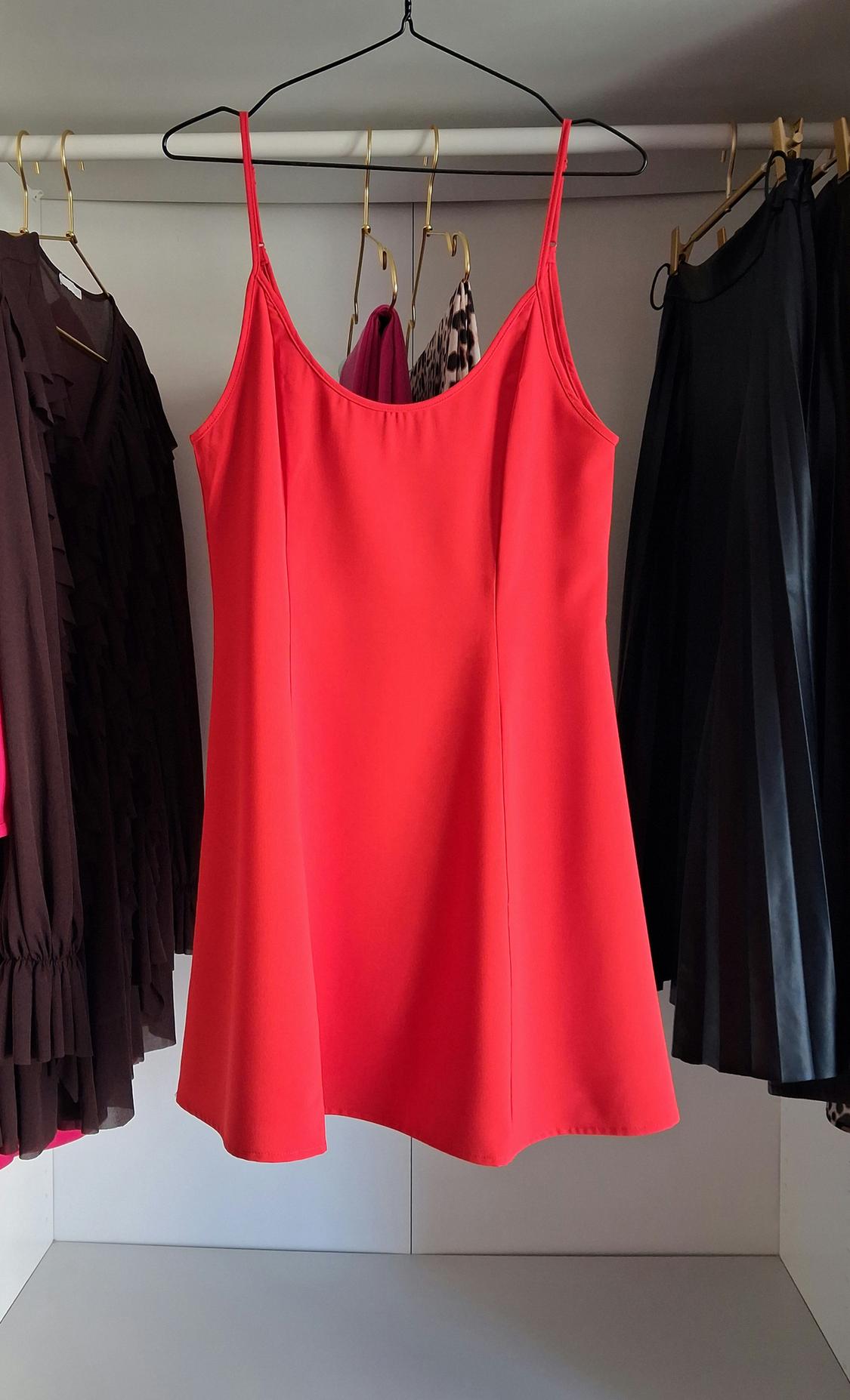 Red strappy dress hanging on a rail, surrounded by other dark garments in an illuminated wardrobe.