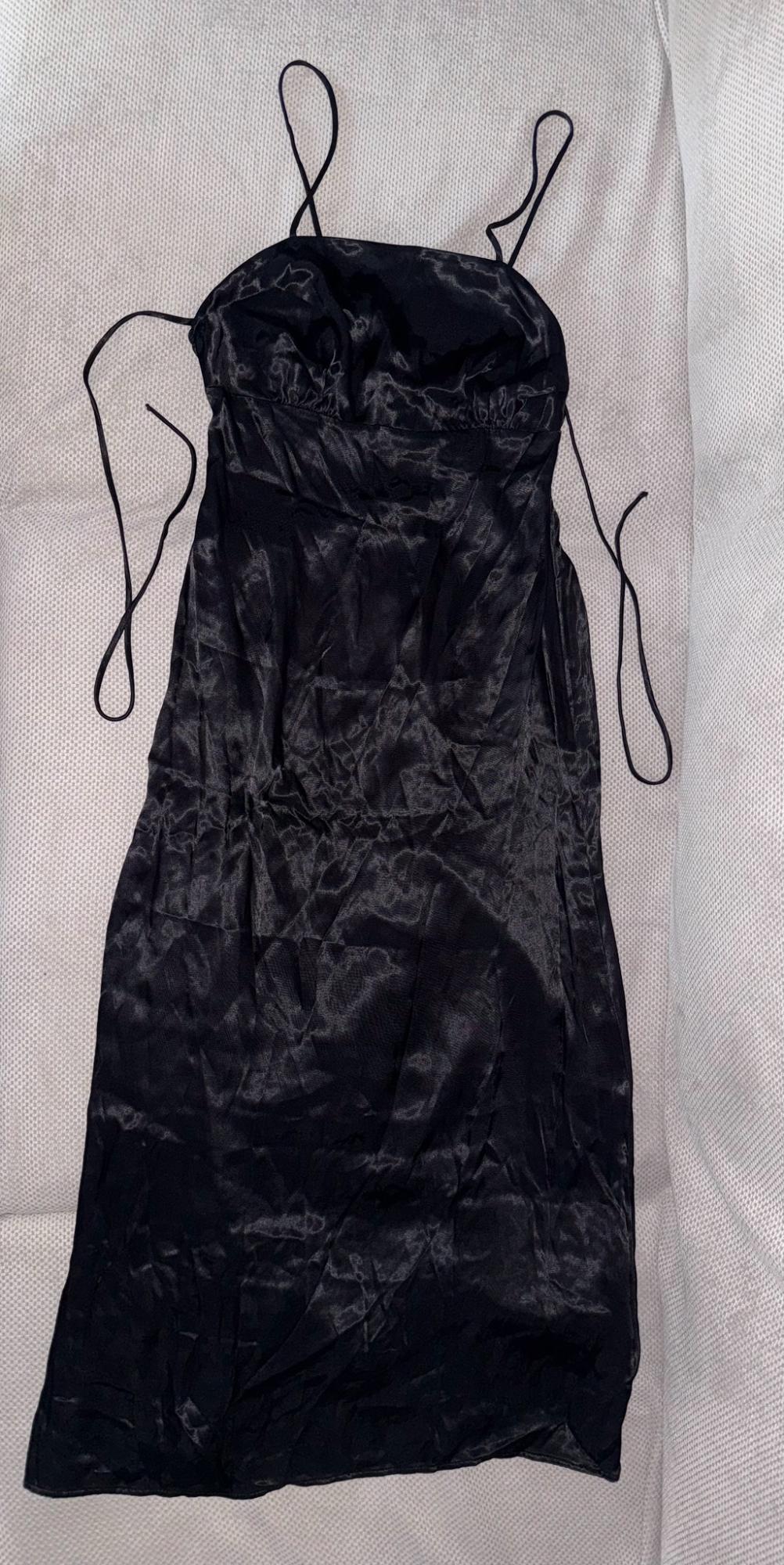 Μαύρο σατέν slip dress με λεπτές τιράντες, απλωμένο πάνω σε μια ανοιχτόχρωμη υφασμάτινη επιφάνεια με υφή. Χαλαρό στυλ.
