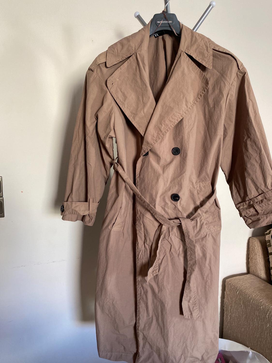 Beige trench coat hanging on a black hanger, with a black belt and buttons, against a white background.