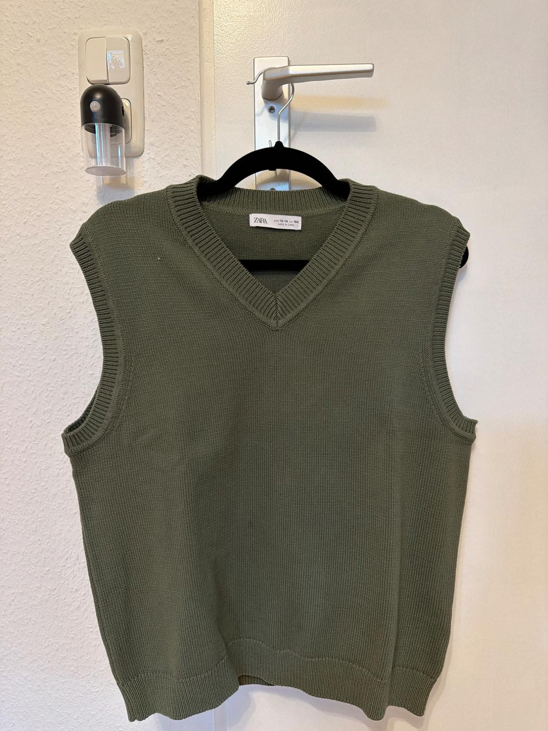 Olive green sleeveless waistcoat hung on a black hanger over a white door. Visible brand label.