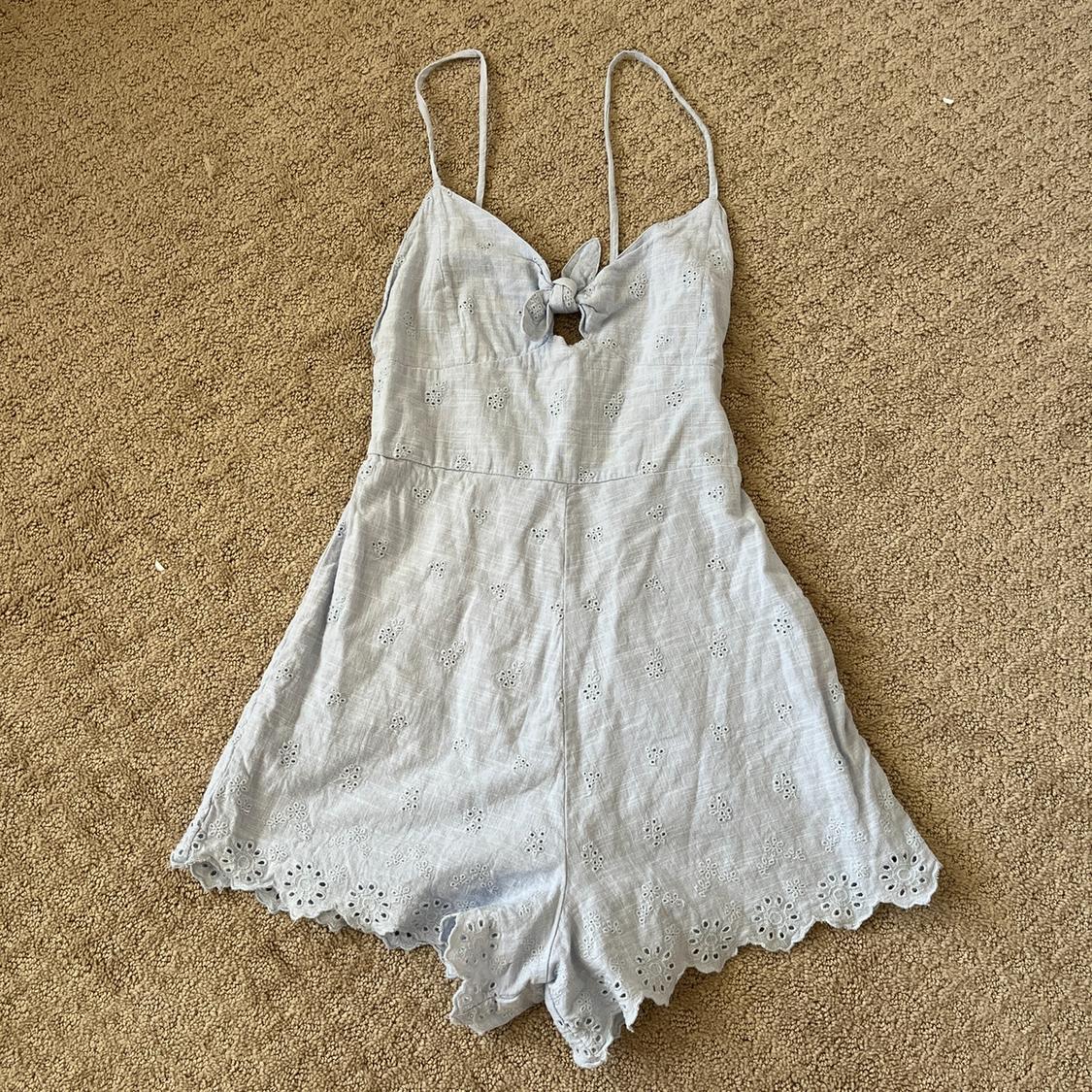 Light blue short romper with spaghetti straps, front knot, and eyelet embroidery. Casual, summery style, on a textured beige rug.
