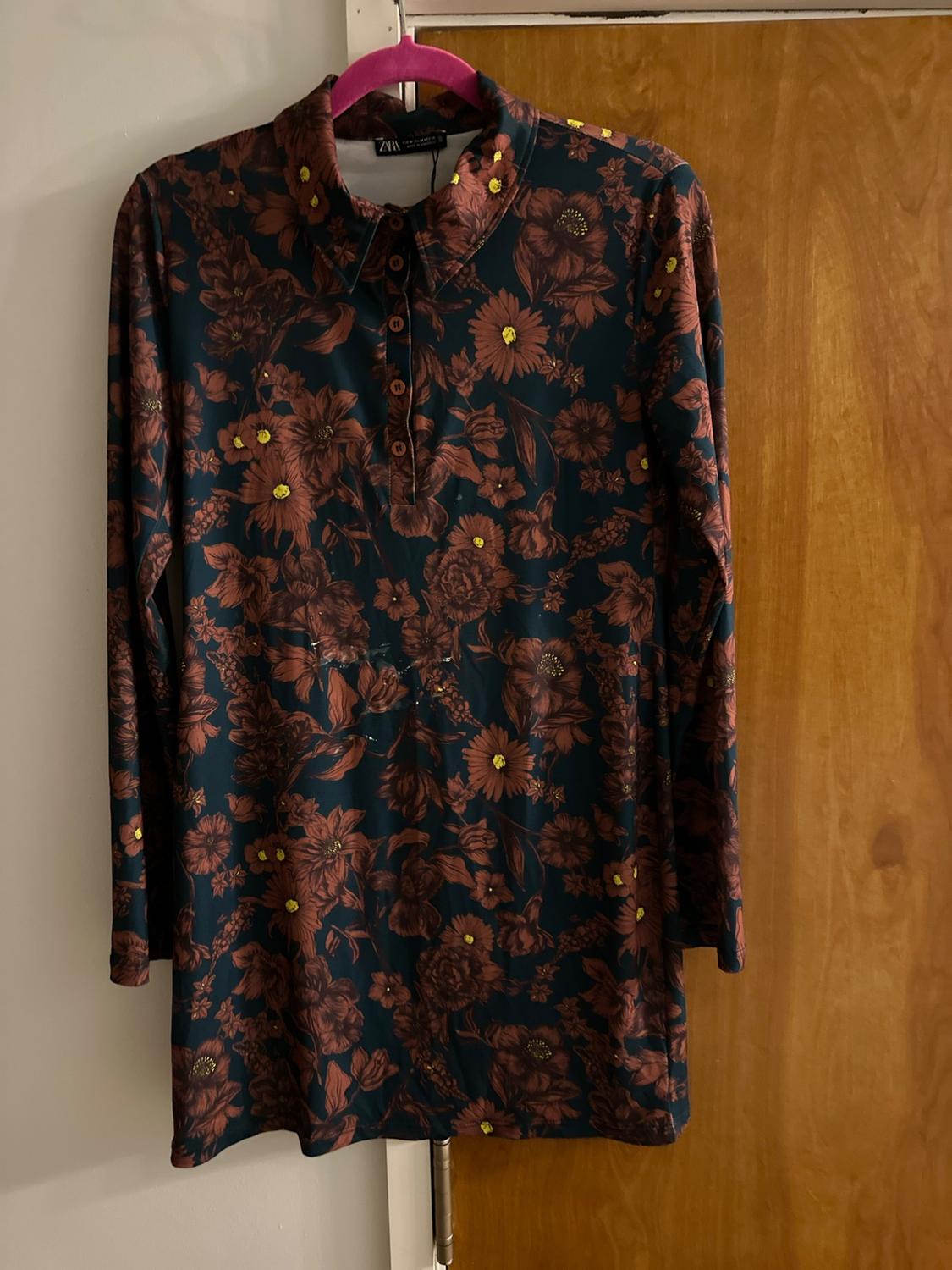Dark dress with a brown and yellow floral print, long sleeves and a button-down collar. ZARA label. Hanging on a pink hanger on a wooden door.