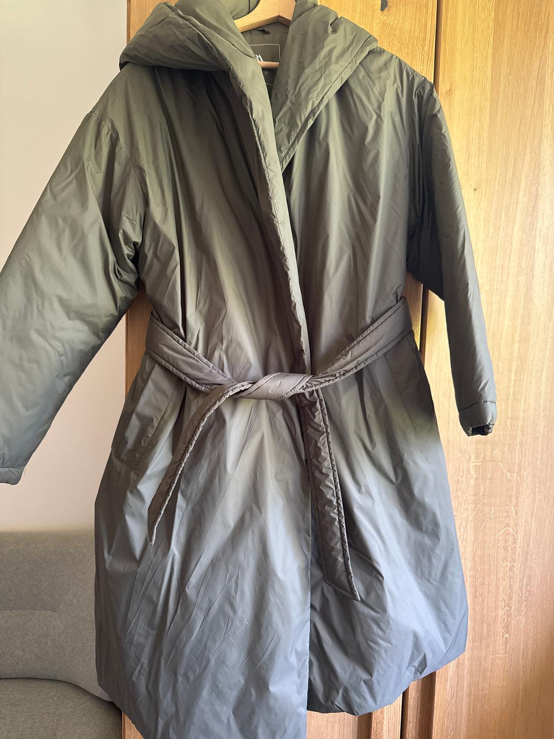 Olive green long padded coat with a hood and belt, hanging on a wooden hanger in front of a light wood wardrobe.