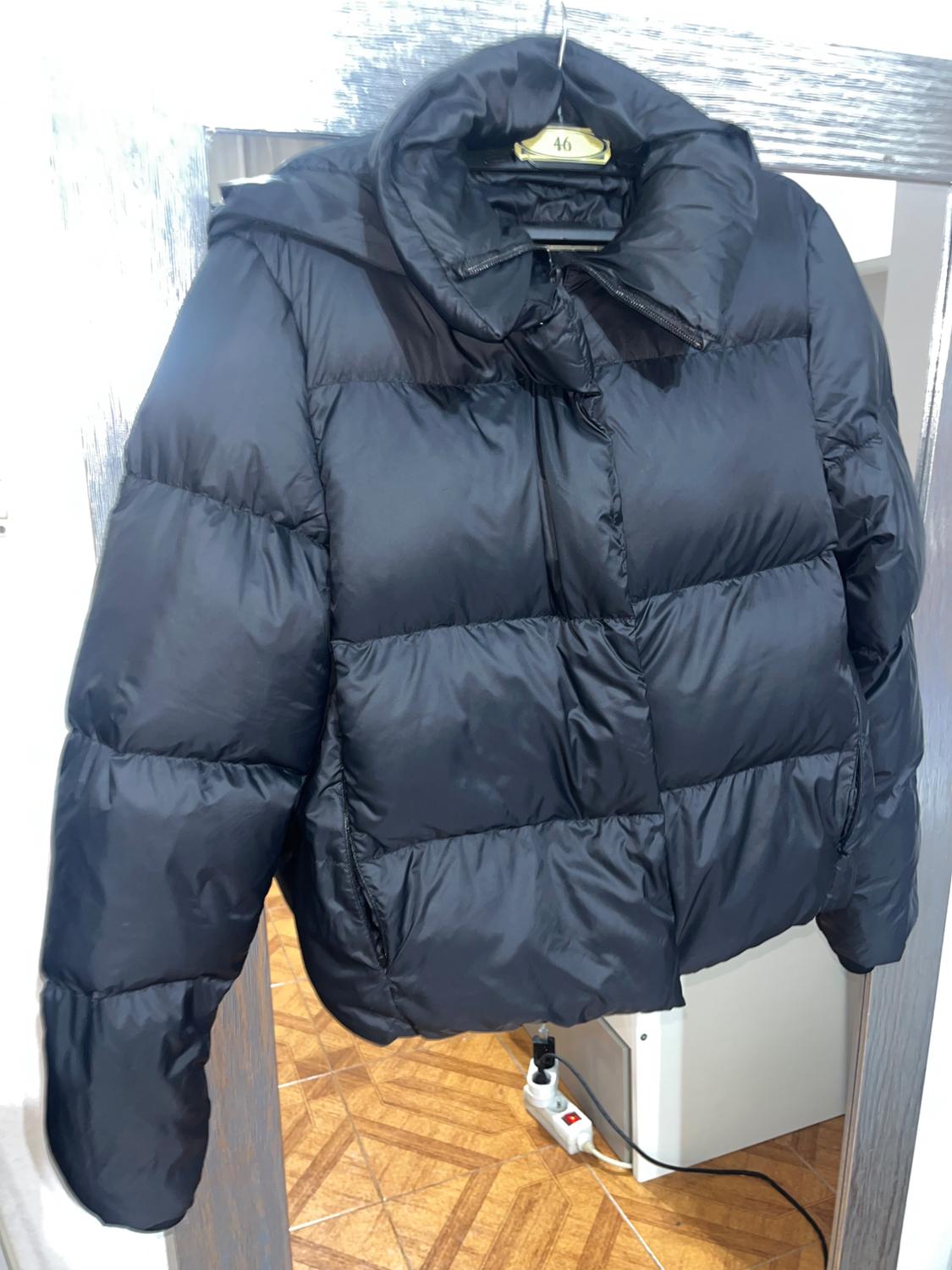 Black padded jacket hanging on a hanger with a size 46 label, against a light wood background and parquet flooring.