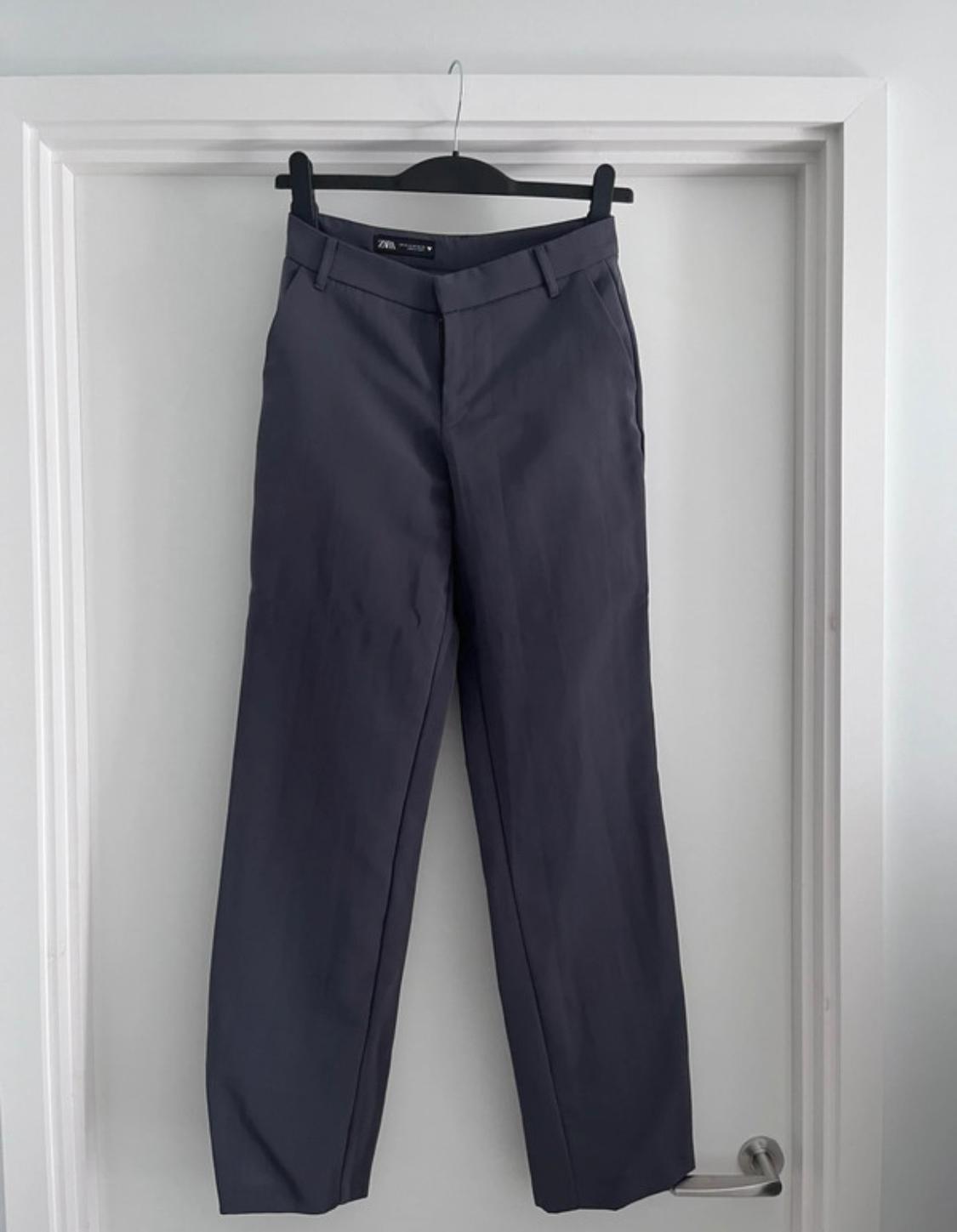 Grey trousers hanging on a black hanger over a white door. Visible label: "Zara Woman".