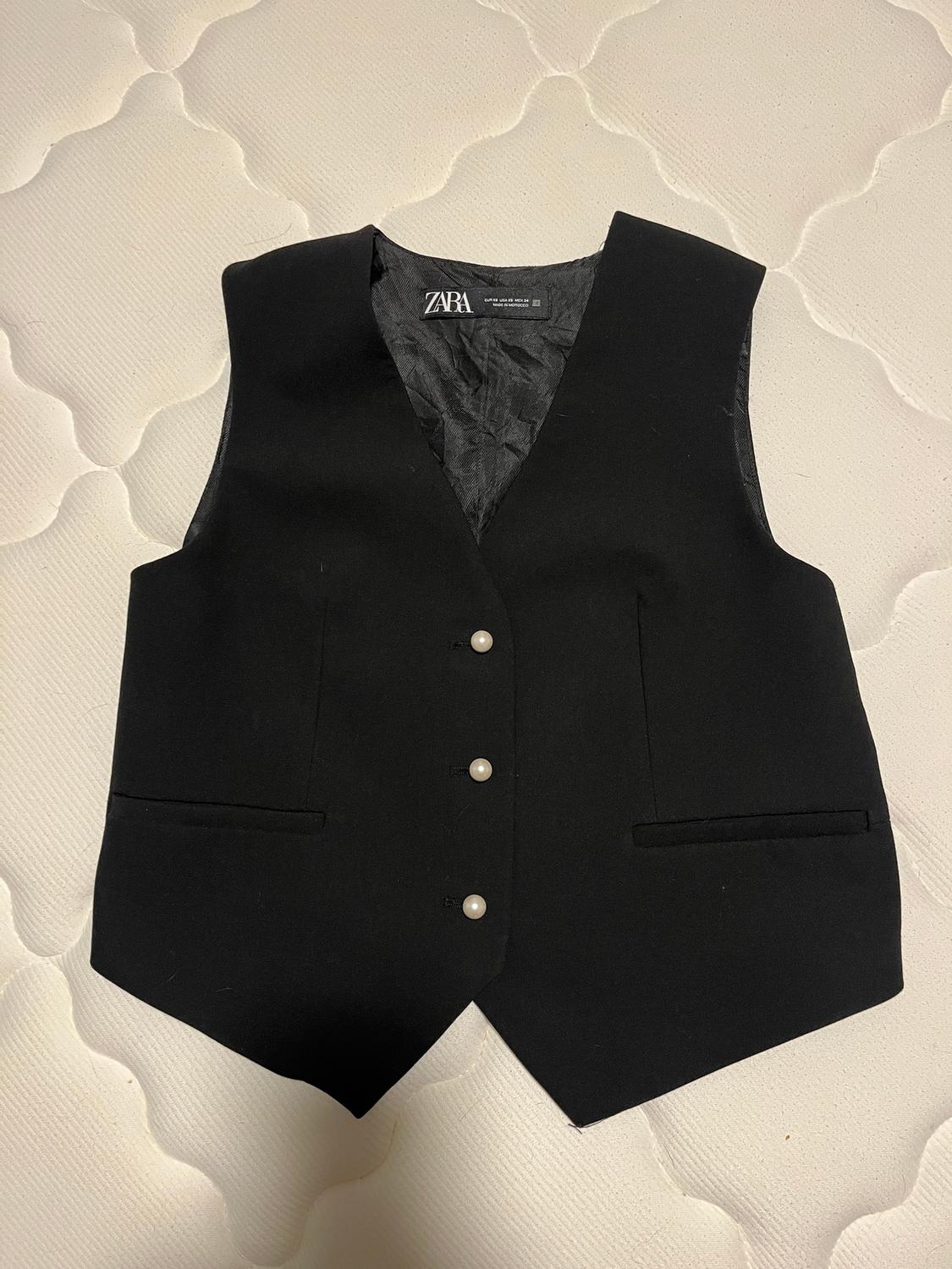 Zara black vest with pearl buttons on a white mattress. Visible tag on the inside of the collar.