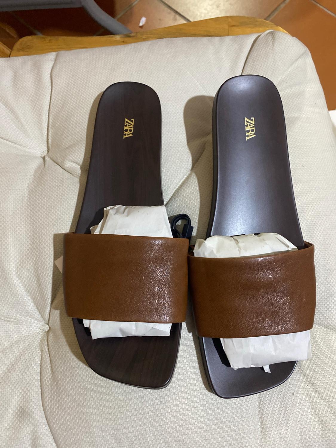 Brown sandals with a wide strap on a light-coloured cushion, stuffed with paper. ZARA text printed on the dark insole.