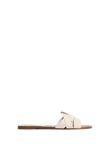 Cream-coloured flat sandal with crossover straps and a brown sole, profile view on a white background.