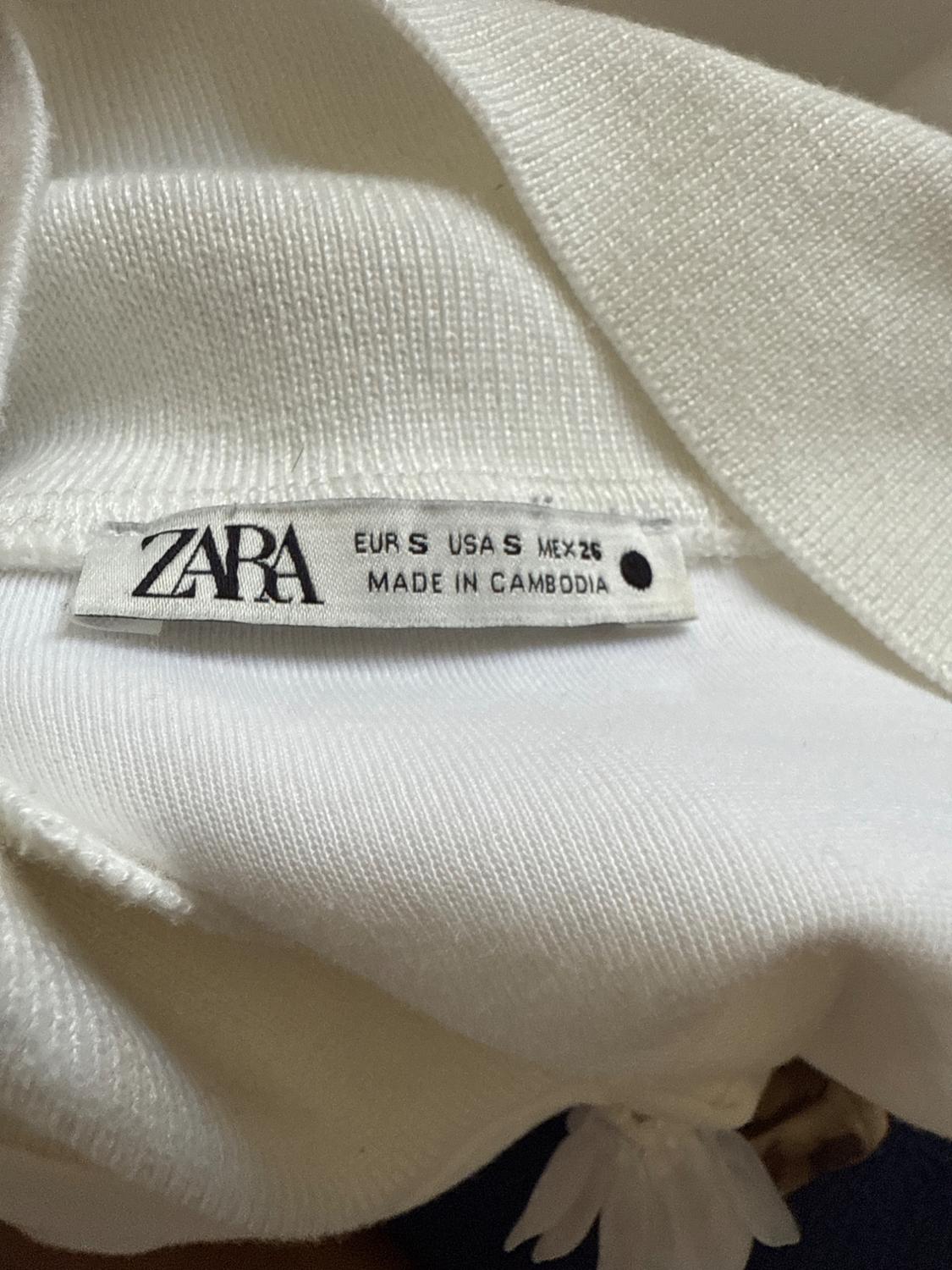 White Zara clothing tag with text: "EUR S USA S MEX 26 MADE IN CAMBODIA" on soft-textured, light-colored fabric.