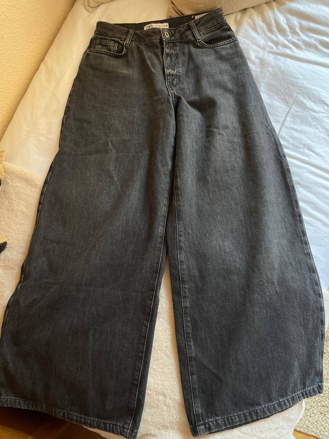 Φαρδύ τζιν παντελόνι από μαύρο denim πάνω σε ένα κρεβάτι με λευκά σεντόνια και μια ορατή ετικέτα στη μέση.