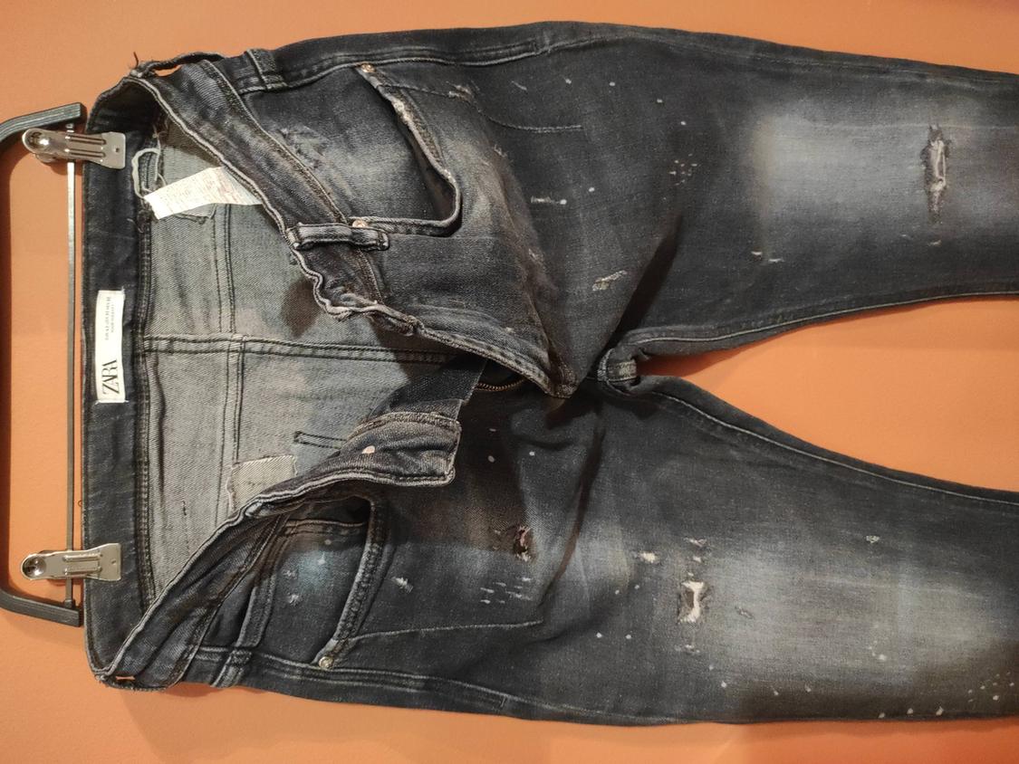 Worn dark denim jeans with ripped details, hanging on a hanger against a brown background. Visible label: "ZARA".