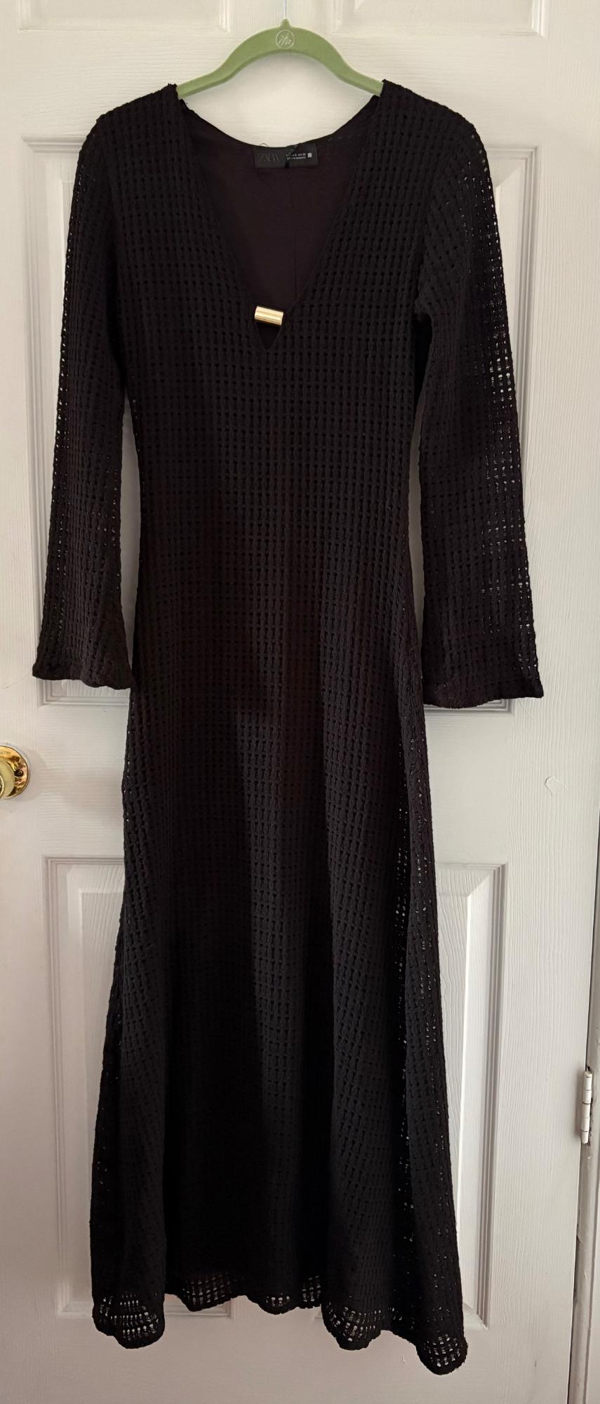 Long-sleeved black dress with openwork fabric and a V-neckline, hanging on a green hanger over a white door.