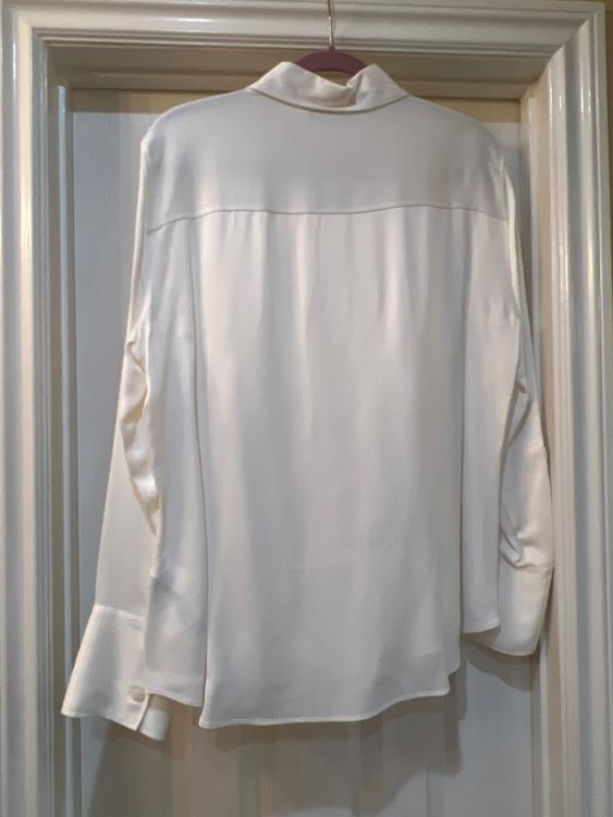 White satin shirt hanging on a hanger inside a door frame, seen from behind, with wide cuffs and a high collar.