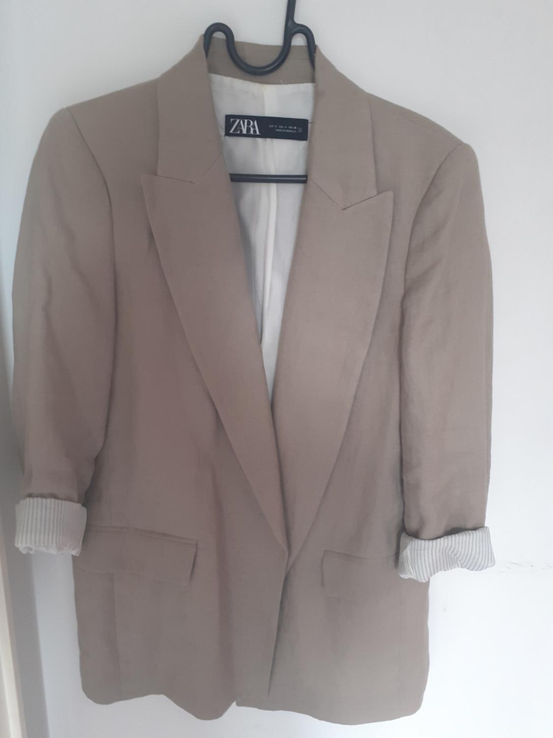 Casual-style beige blazer on a hanger against a white wall. Sleeves rolled up with a striped lining. Label with the text Zara.