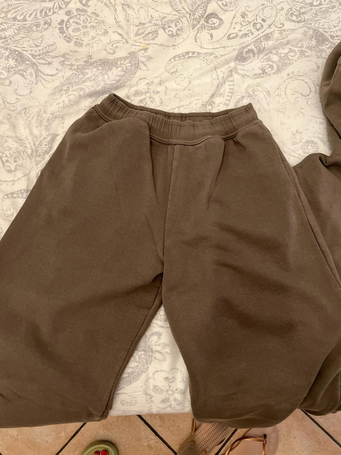 Brown tracksuit bottoms on a bed with patterned sheets. Casual and relaxed style. Footwear and the floor are visible at the bottom.