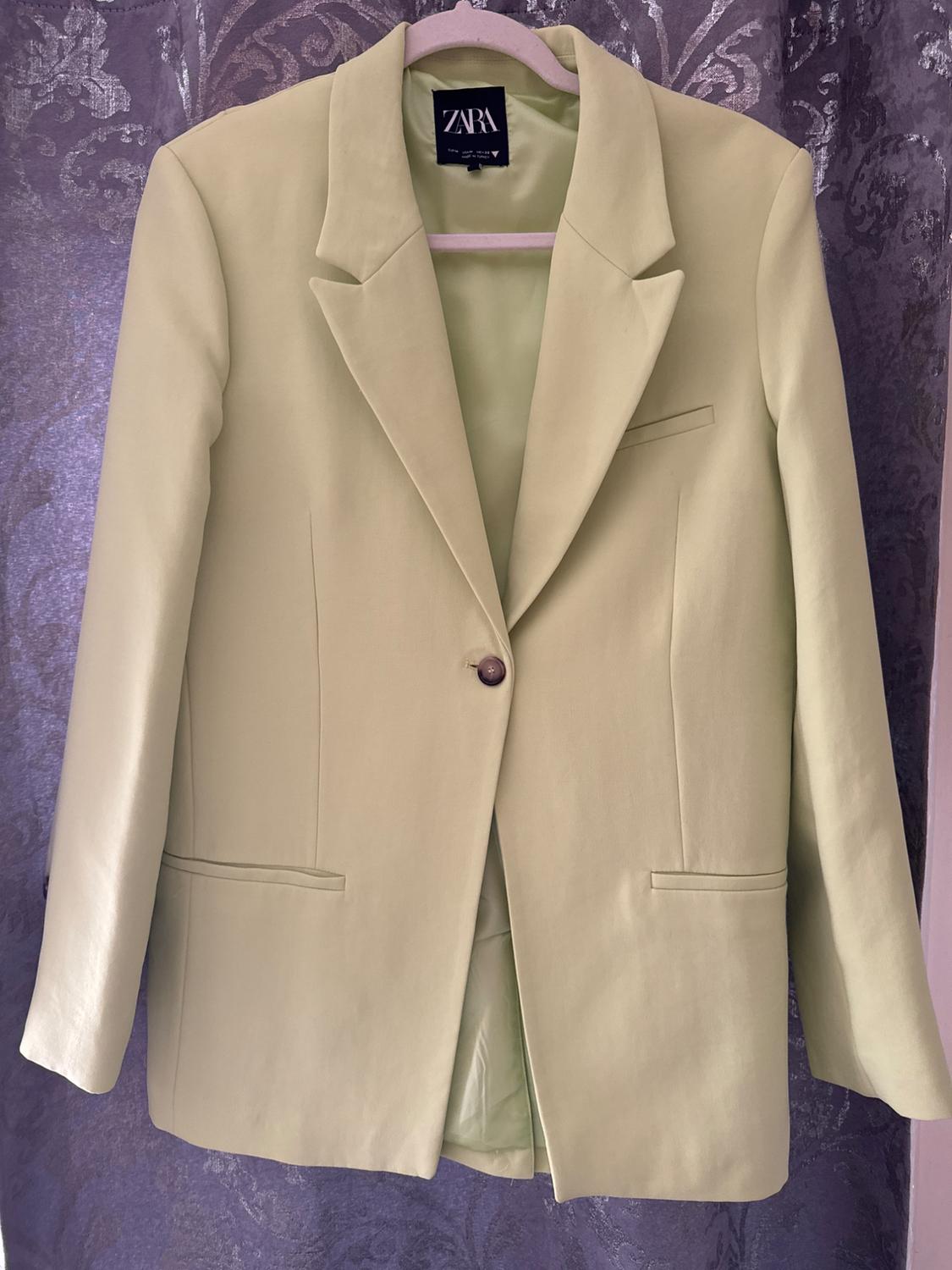 Light green jacket hanging on a hanger, with a front button and a visible Zara tag, against a floral fabric background.
