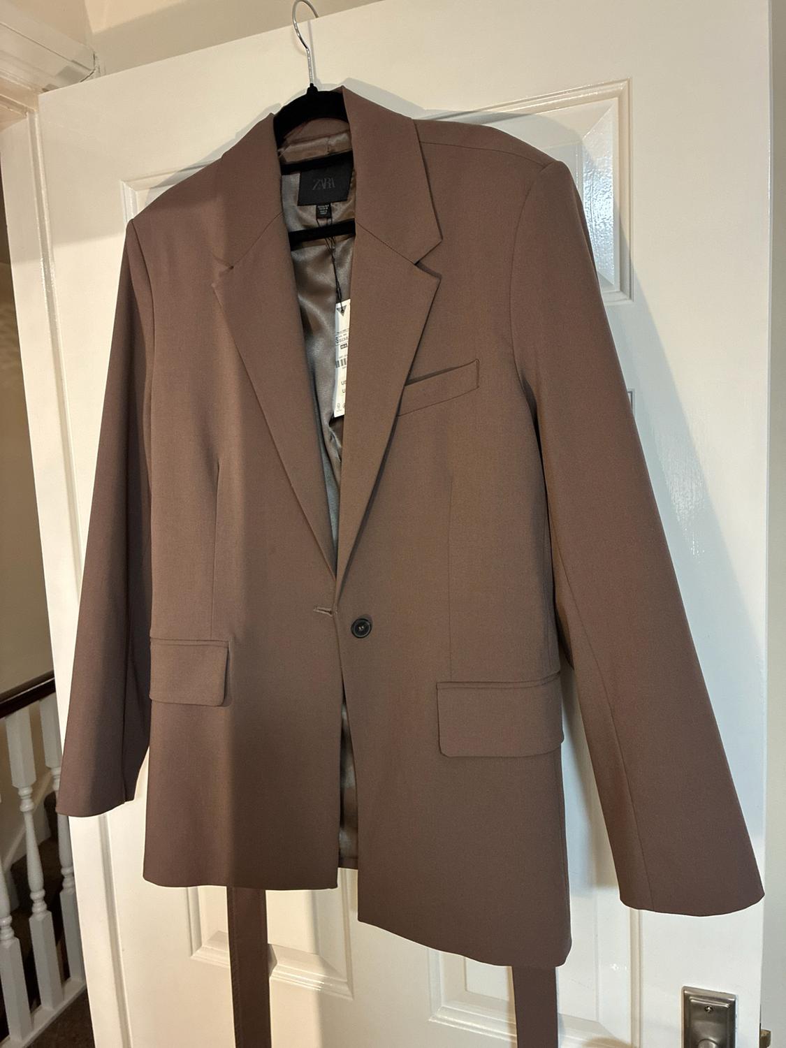 Brown blazer hanging on a white door. Interior label with ZARA text. Features one button, flap pockets and a belt.