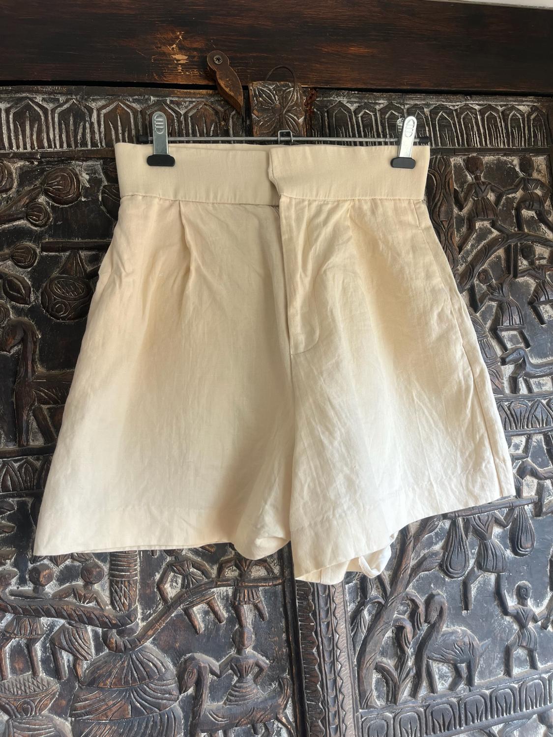 Beige pleated shorts hanging on a hanger against a dark carved wood background. Rustic and relaxed style.