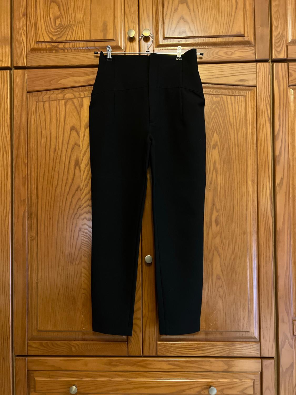 High-waisted black tailored trousers, hanging in a wooden wardrobe. Centred composition that conveys a relaxed and homely atmosphere.