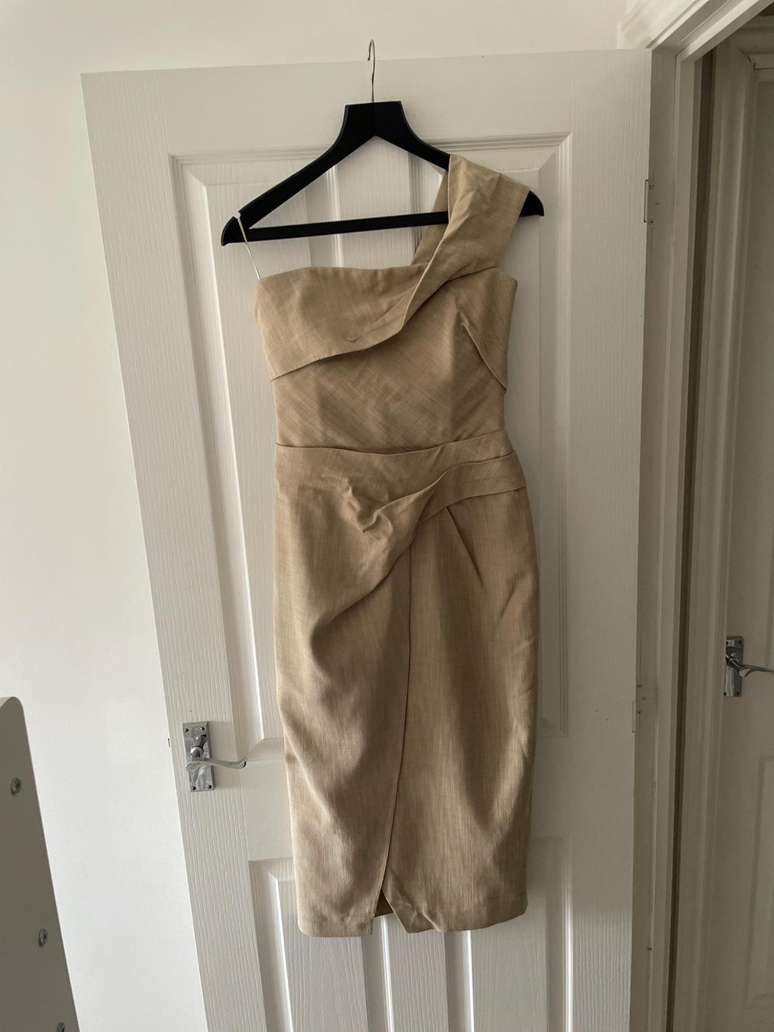 Beige one-shoulder dress with draped detail, hanging on a black hanger on a white interior door.