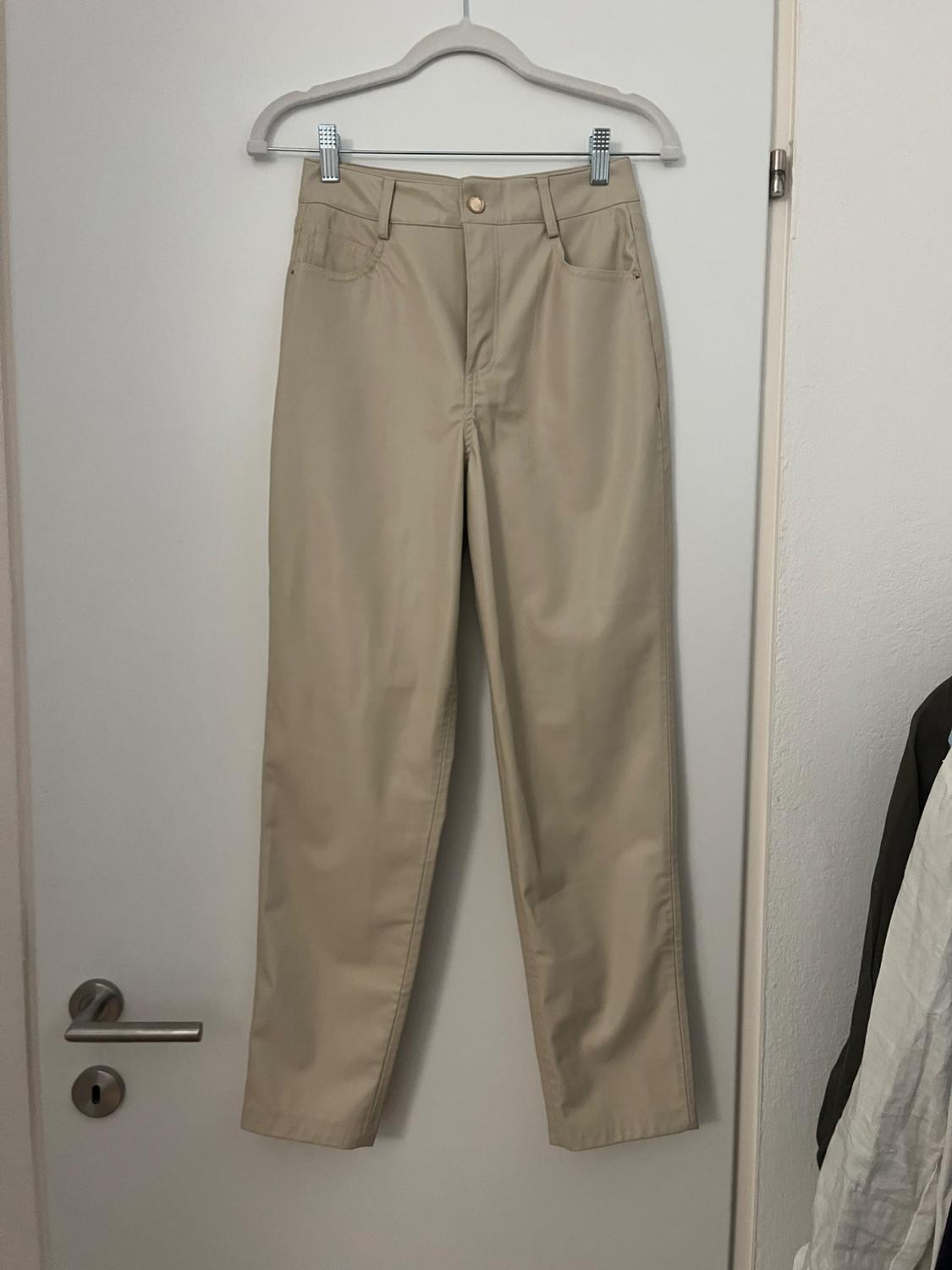 Beige trousers hanging on a white hanger over a white door. Classic design with pockets and a gold button.